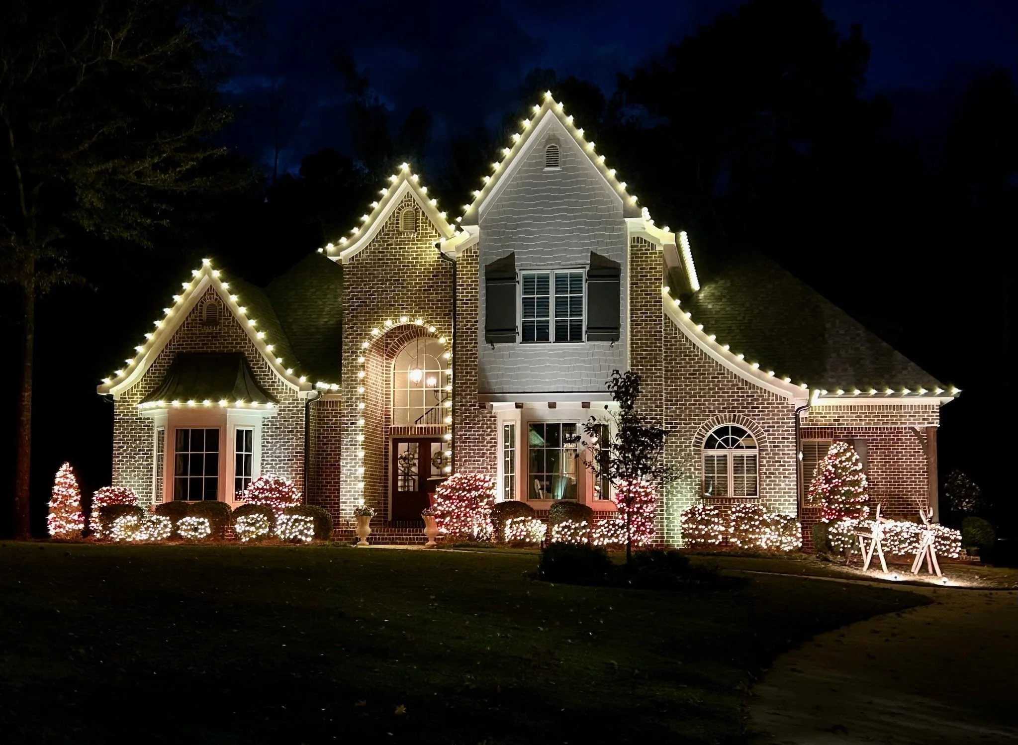 A large brick house decorated with white Christmas lights outlining the roof and windows. The front yard features illuminated bushes and trees, with some Christmas decorations visible near the entrance.