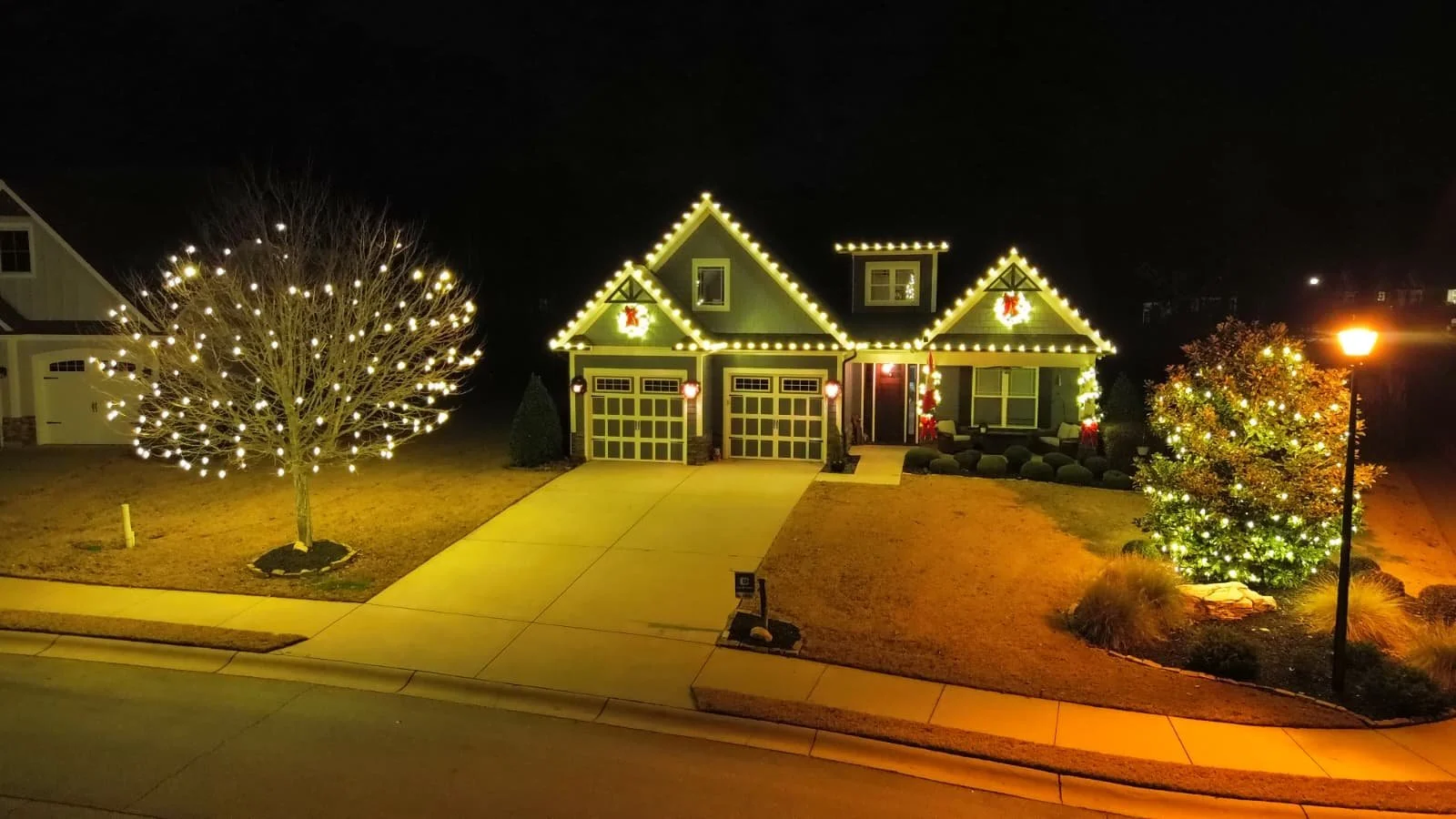 A house decorated with Christmas lights at night, with a porch, two trees with lights, and a streetlamp nearby.