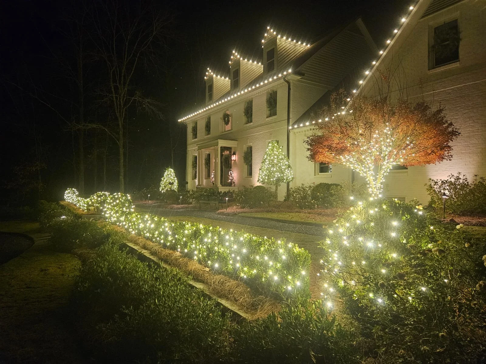 A large house decorated with Christmas lights at night. The house has string lights outlining the roof, and the yard is decorated with illuminated trees and bushes.