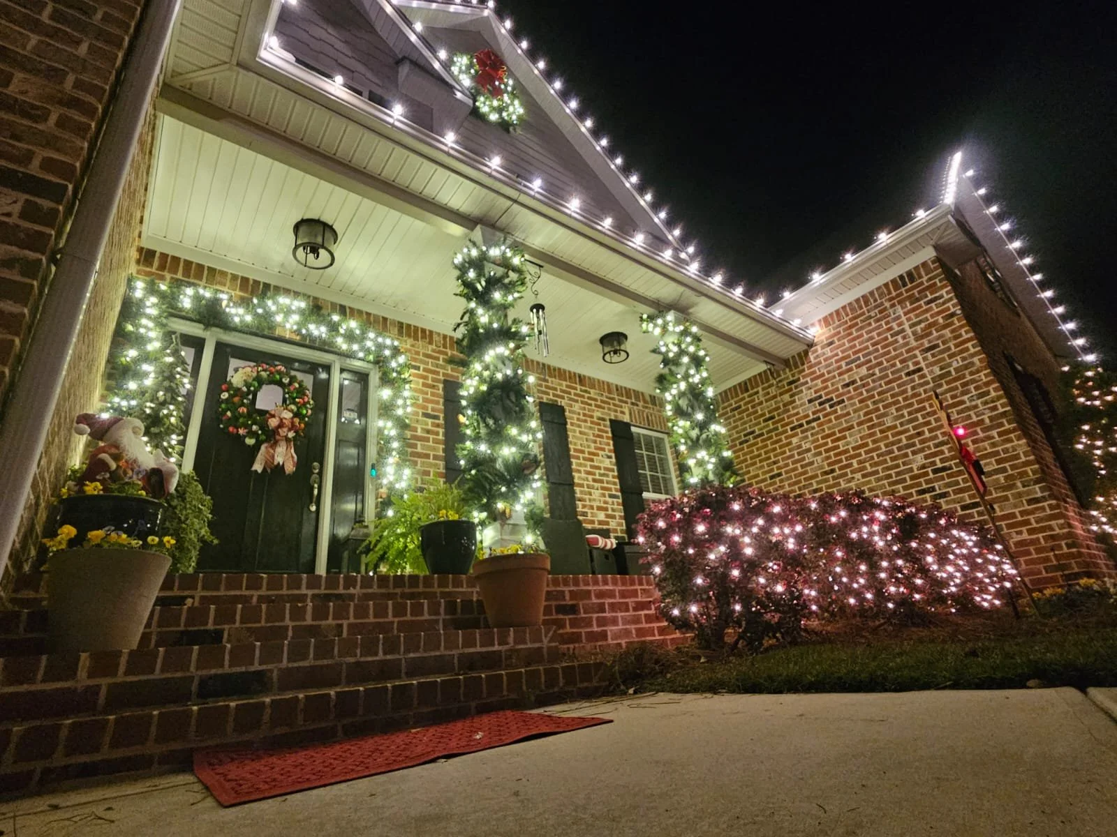 A front porch decorated for Christmas with white string lights, trees wrapped in lights, a wreath on the door, and potted plants, including a Santa Claus figure, under a dark night sky.