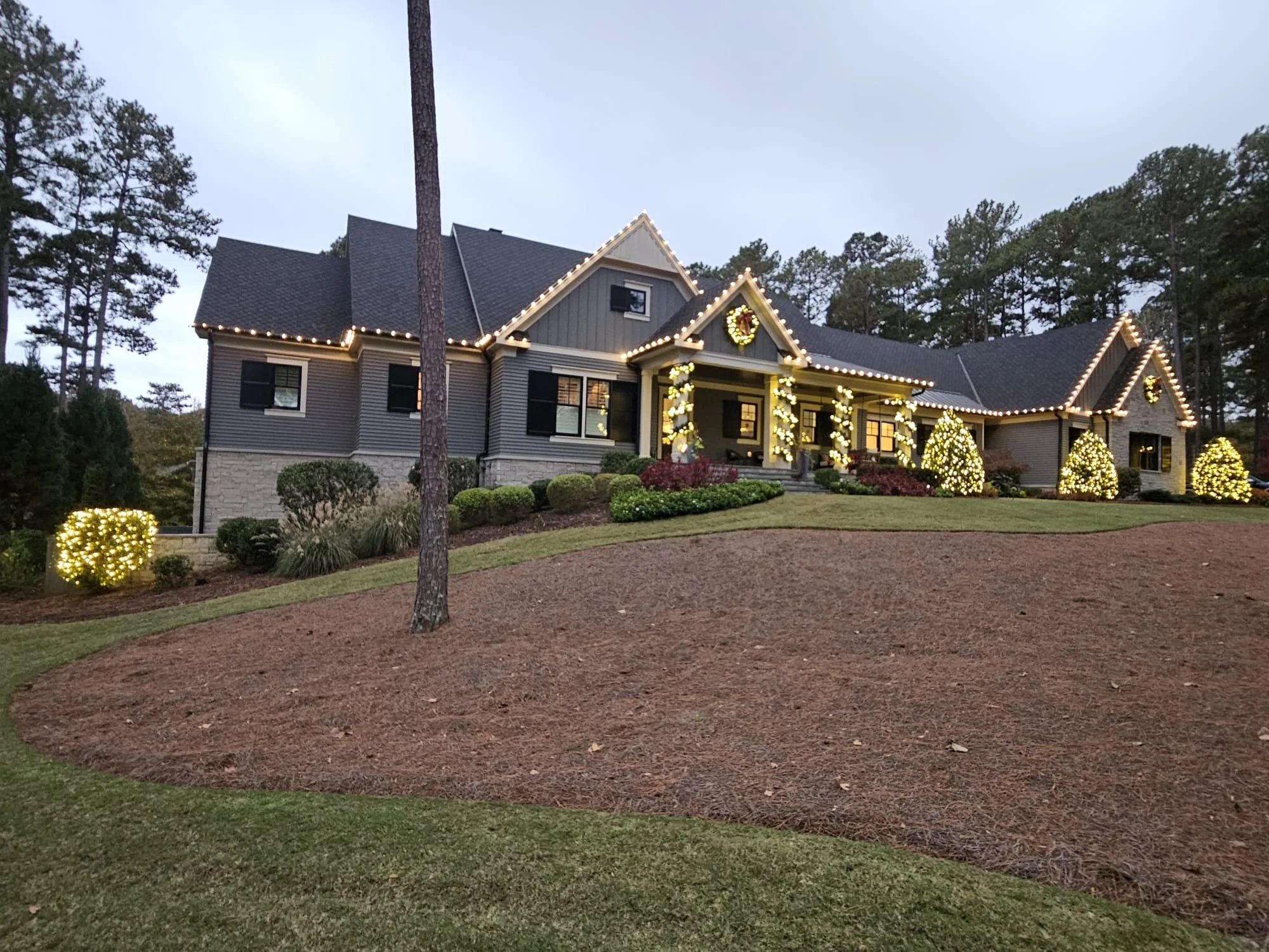 A house decorated for Christmas with string lights along the roofline and windows, two wreaths, and lit bushes in the yard, surrounded by trees and a partly cloudy sky.