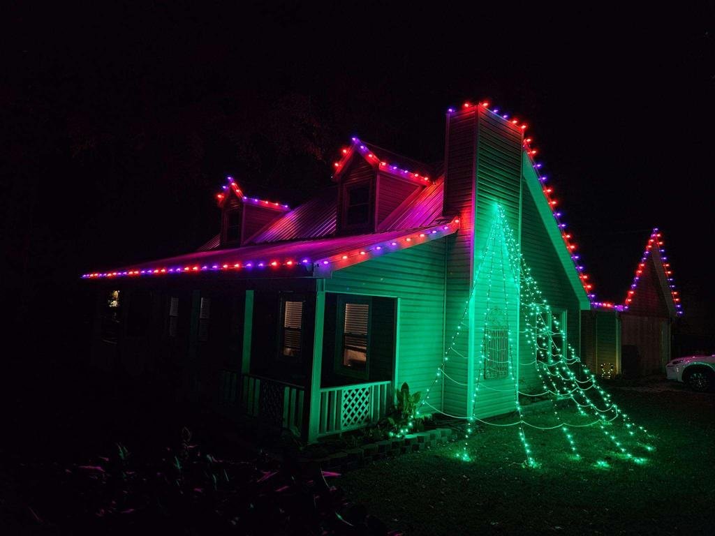 House decorated with colorful holiday lights at night, featuring green and red lights on the roof and green lights on a tree-shaped display on the side.