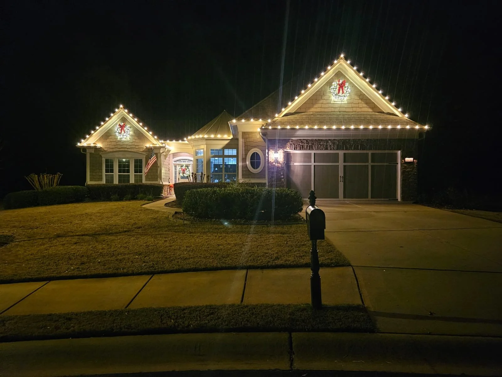 A well-lit house decorated with Christmas holiday lights and wreaths at night, with a front yard, driveway, and a mailbox.