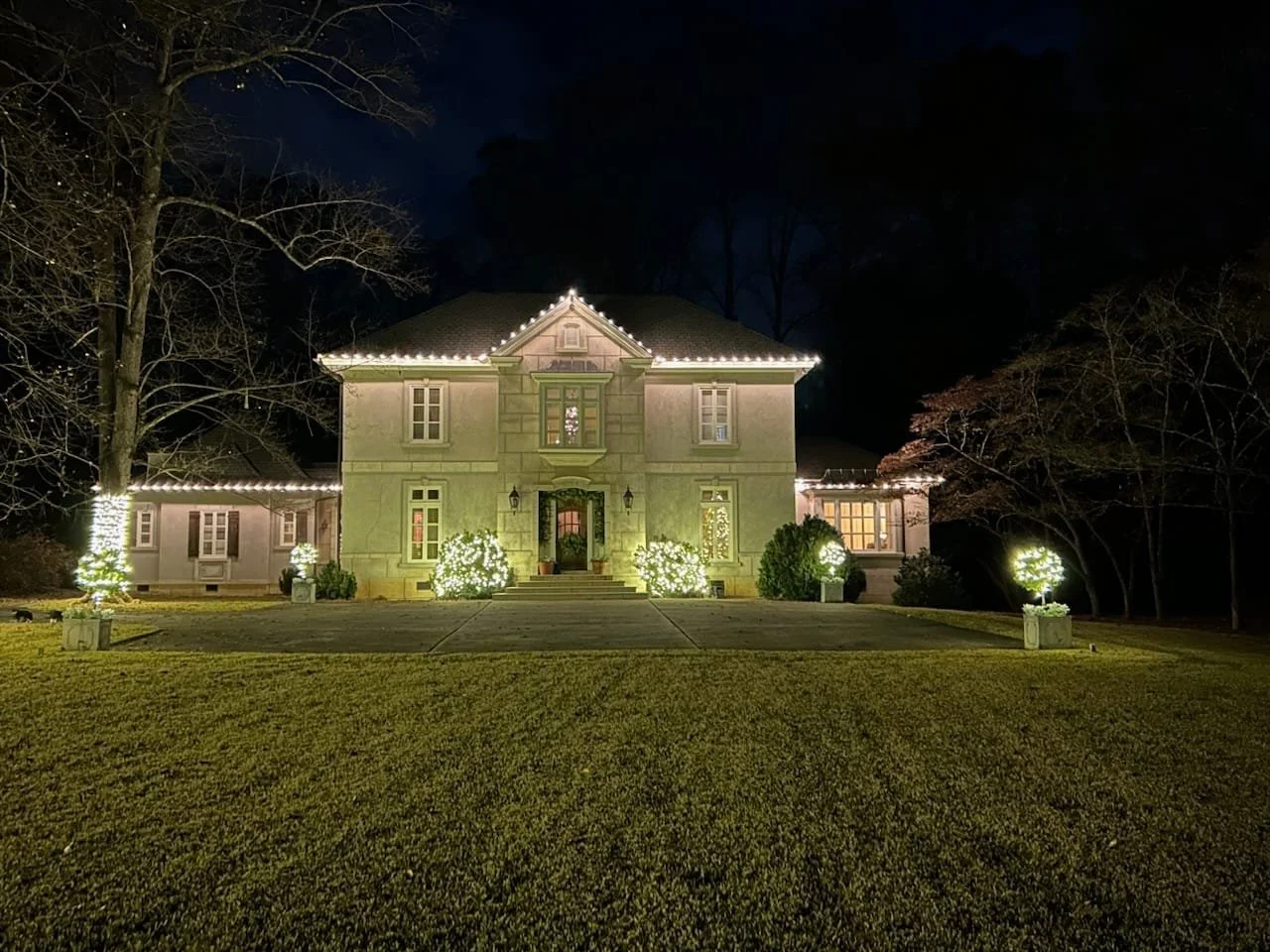 A large house decorated with Christmas lights at night, with Christmas trees and bushes illuminated outside.