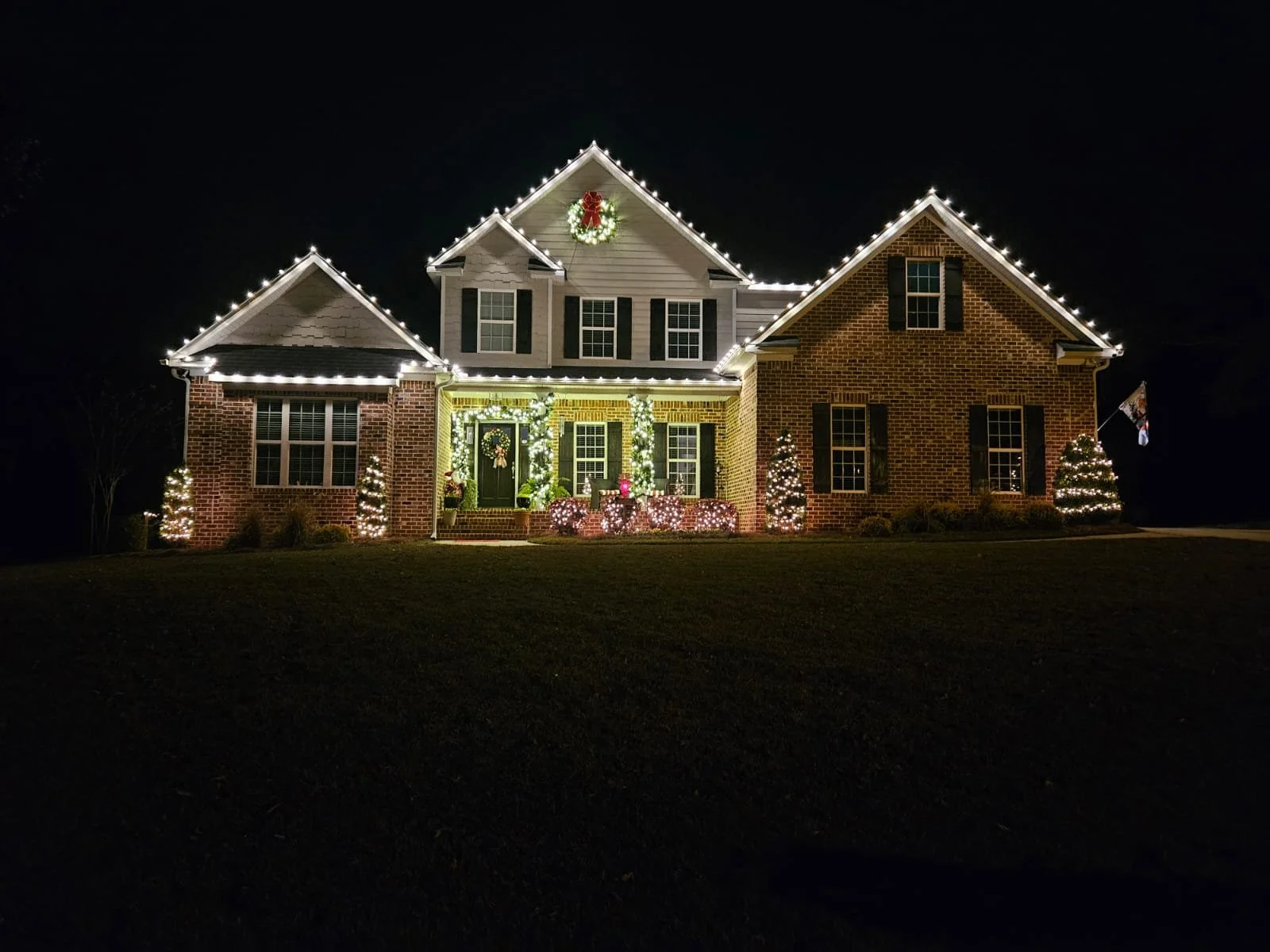 House decorated with Christmas lights and ornaments at night, with a wreath on the porch and small trees wrapped in lights.