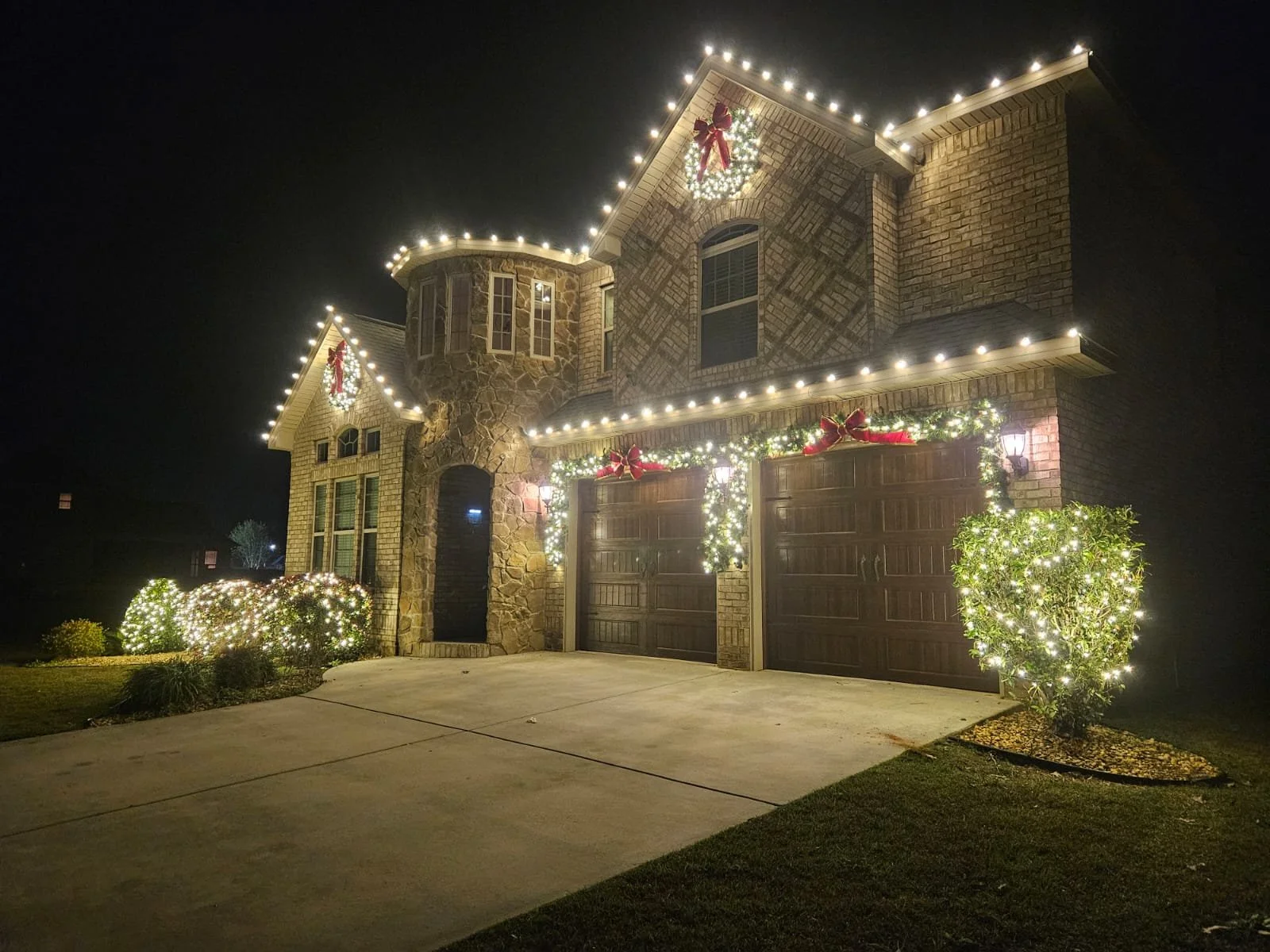 A two-story house decorated with Christmas lights and wreaths, featuring a stone and brick exterior, with bright outdoor lighting, at night.