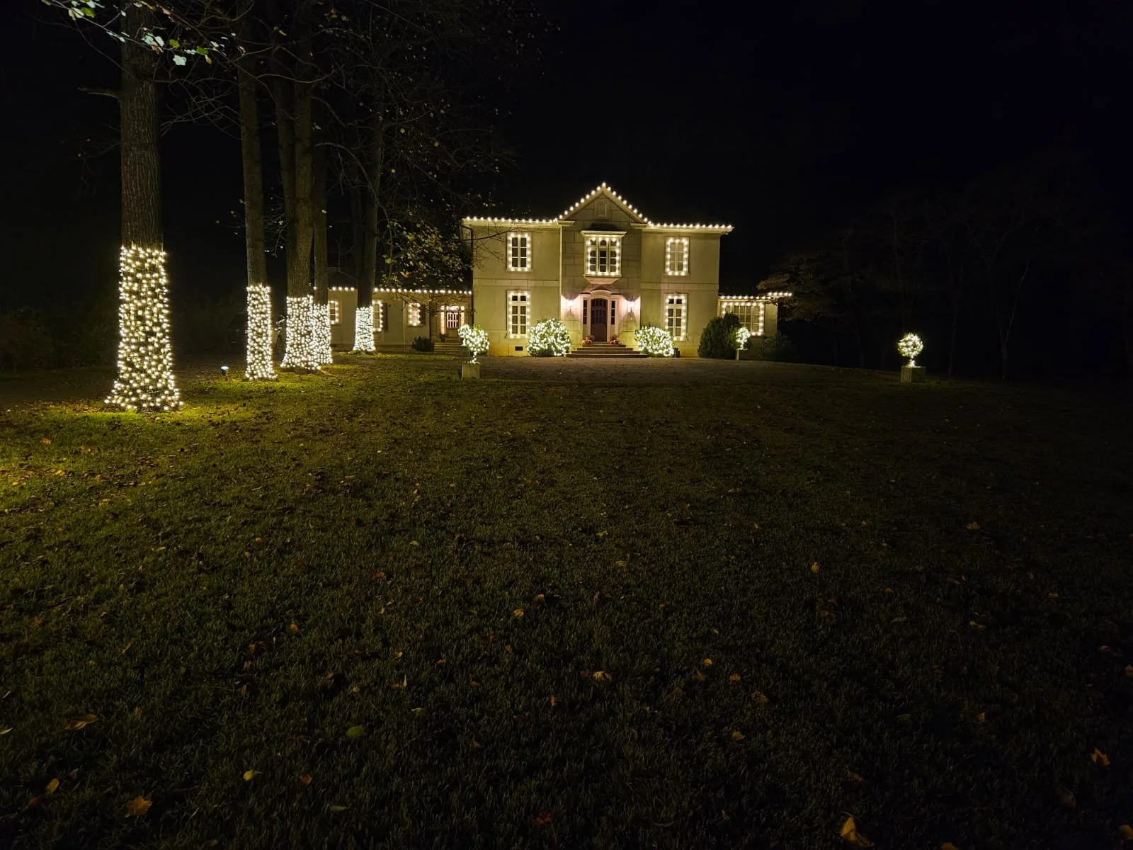 Night view of a large house decorated with Christmas lights outlining windows, doors, and roof. Trees in front of the house have wrapped string lights at their bases, and bushes are lit with spotlights, creating a festive holiday scene.
