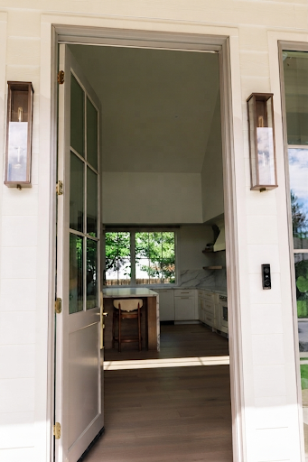 Open front door revealing a kitchen with white cabinets, windows showing green trees outside, a small island, and light wood flooring.