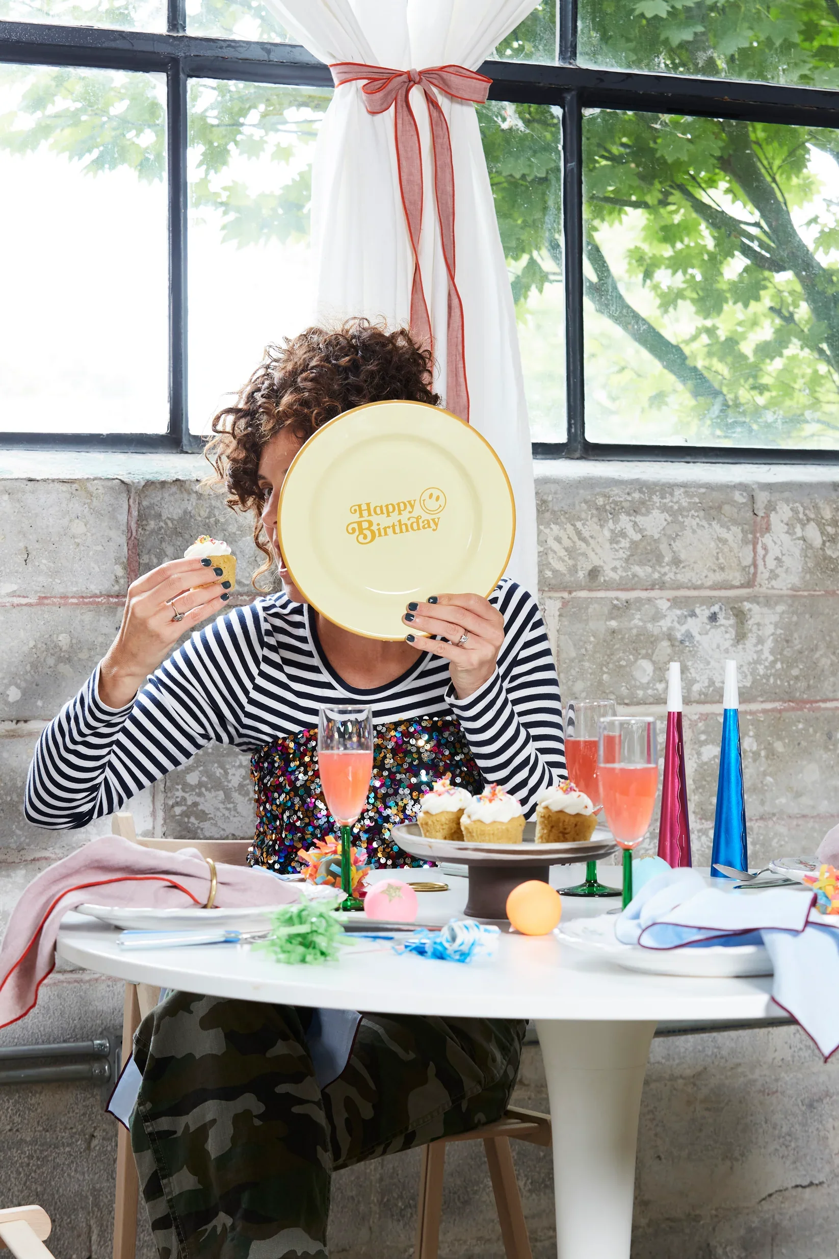 A woman with curly hair in a striped shirt, sitting at a table, holding a plate covering her face and a cupcake in her other hand. The table is decorated with cupcakes, pink drinks, and party decorations, with a window and white curtains in the background.