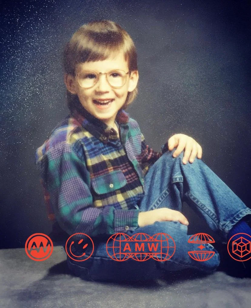 A young boy with glasses and brown hair, smiling, sitting on a black surface against a dark background, wearing a colorful plaid shirt and jeans.
