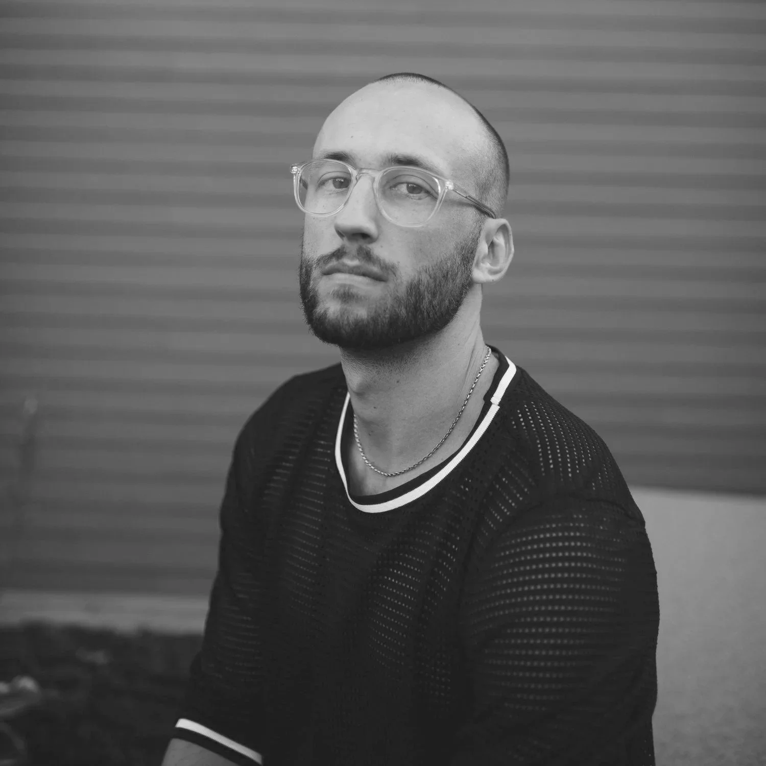 Black and white portrait of a man with a beard and glasses, wearing a black shirt with striped trim and jewelry, standing against a horizontally striped background.