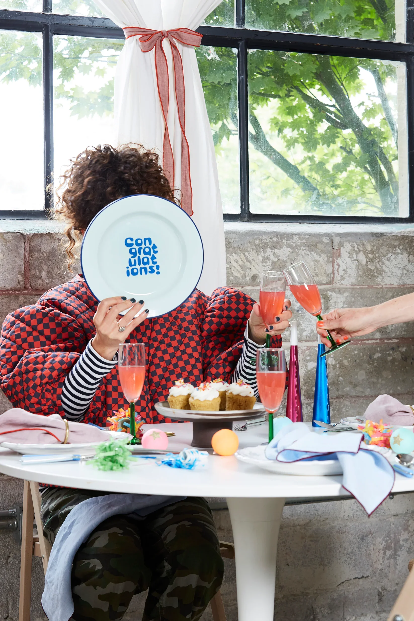 A woman with curly hair sitting at a decorated party table, holding a white plate with the words "Congratulations!" on it, partially covering her face. The table has cupcakes, colorful party hats, balloons, and glasses of pink sparkling drink. There are colorful party blowers and decorations on the table. The background features a window with green trees outside and white curtains tied with a red ribbon.