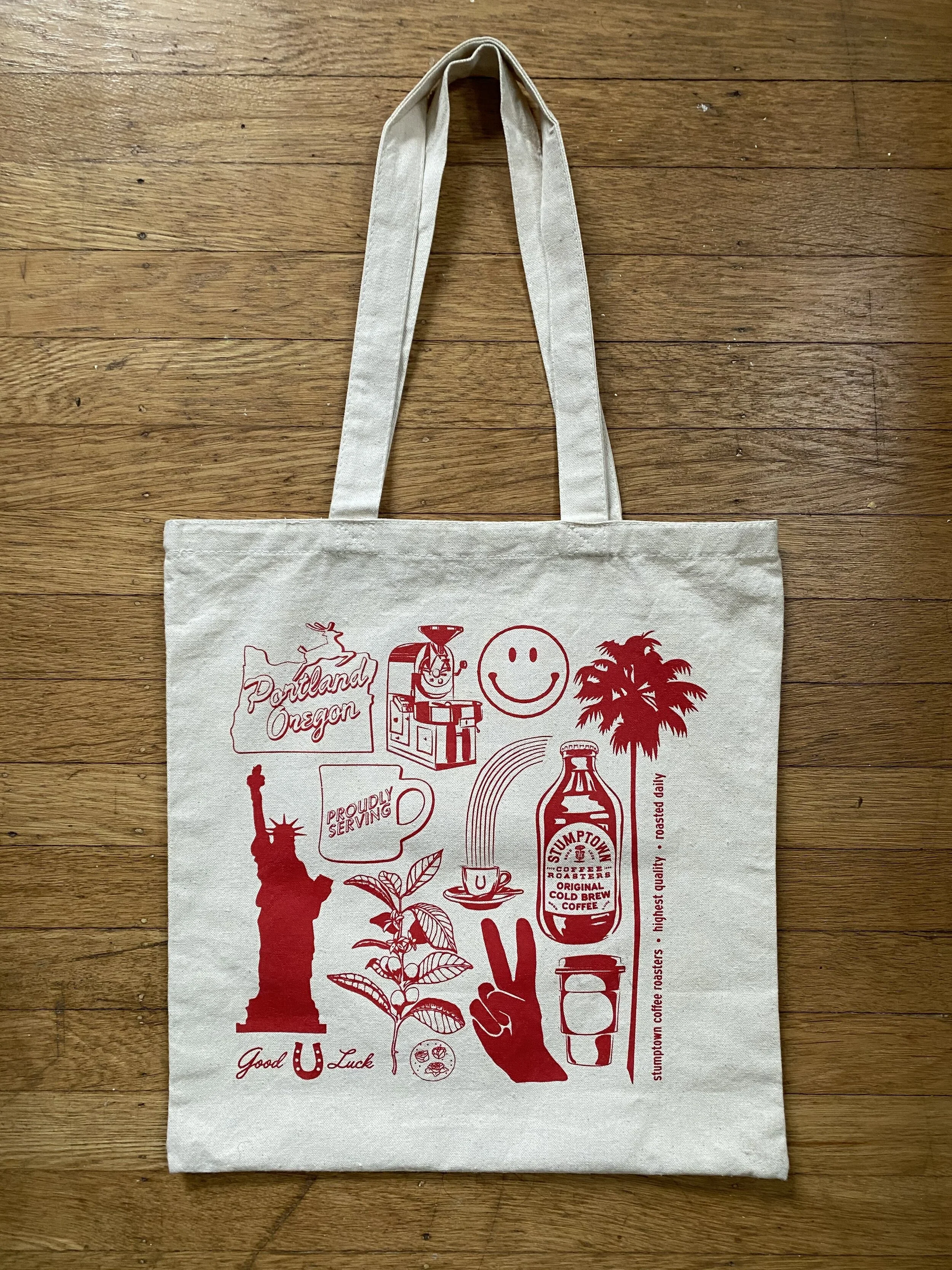 A beige tote bag with red illustrations and text, featuring images of the Statue of Liberty, a coffee bottle, a smiling face, and various other icons, lying on a wooden floor.