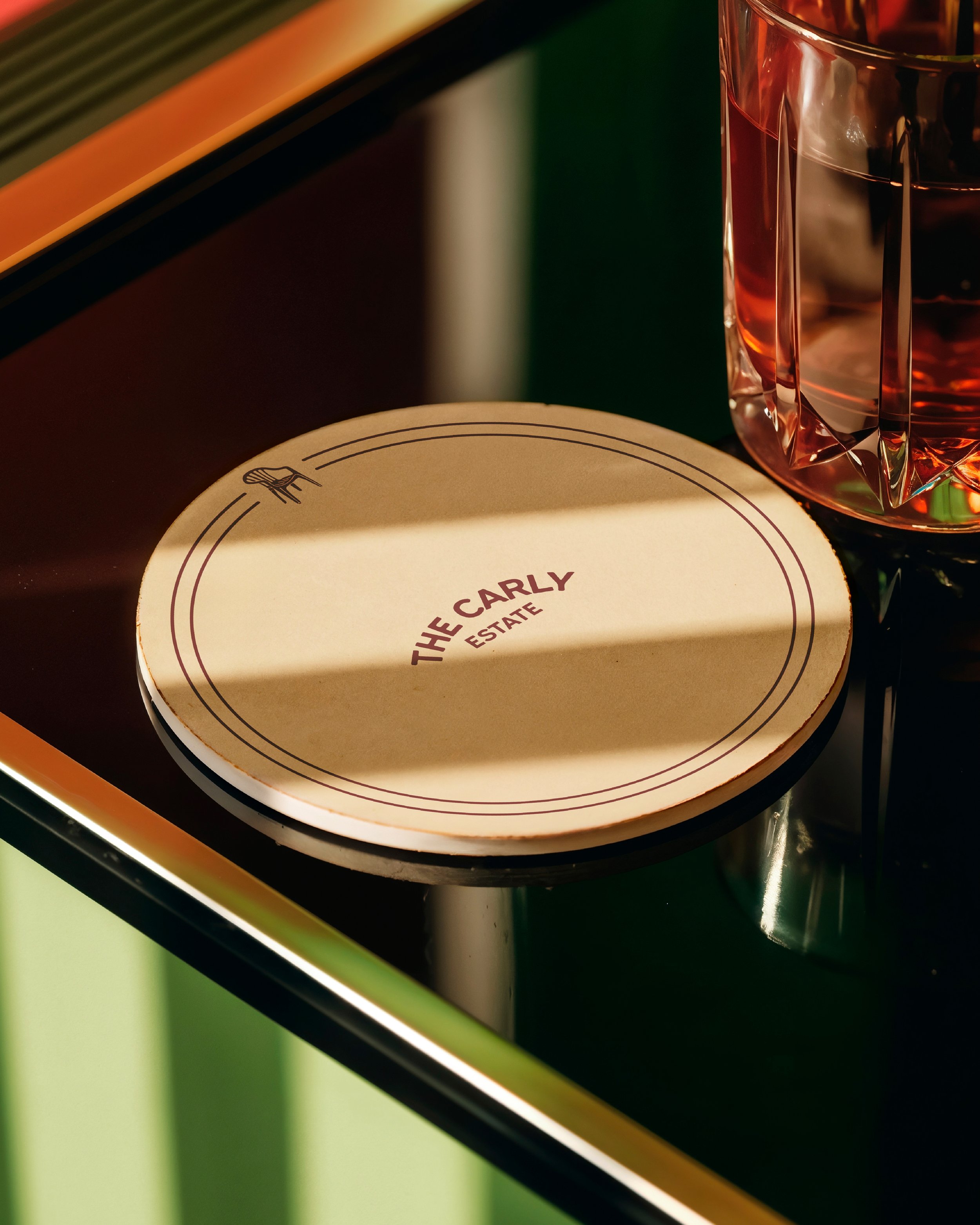 Close-up of a round pizza box with a beige lid partial logo reading "The Carly" and a wine glass with a reddish drink on a reflective black surface.