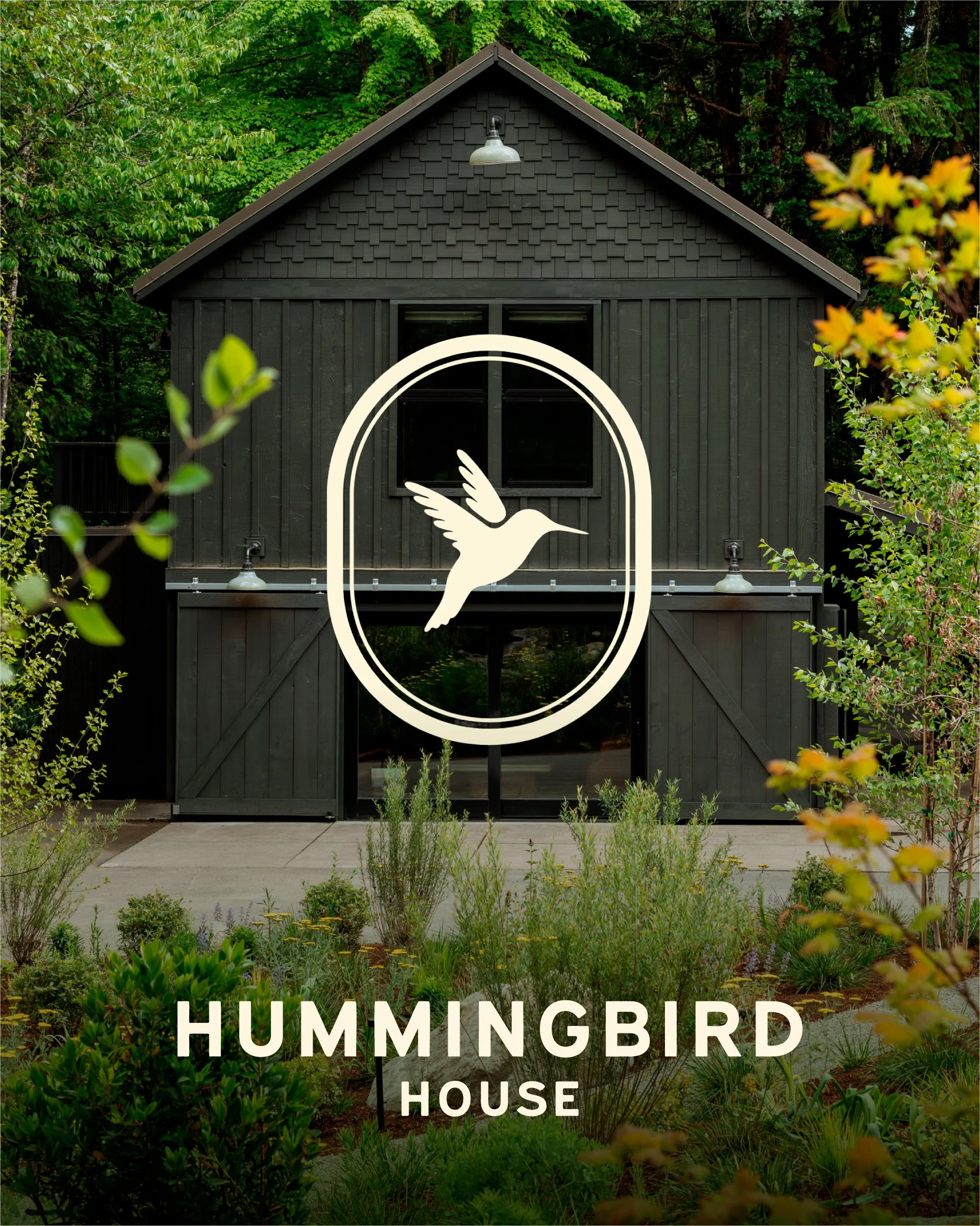 A dark-colored barn with a pitched roof, surrounded by lush green trees and plants. The barn features a large white sign with a hummingbird graphic and the text 'HUMMINGBIRD HOUSE' beneath it.