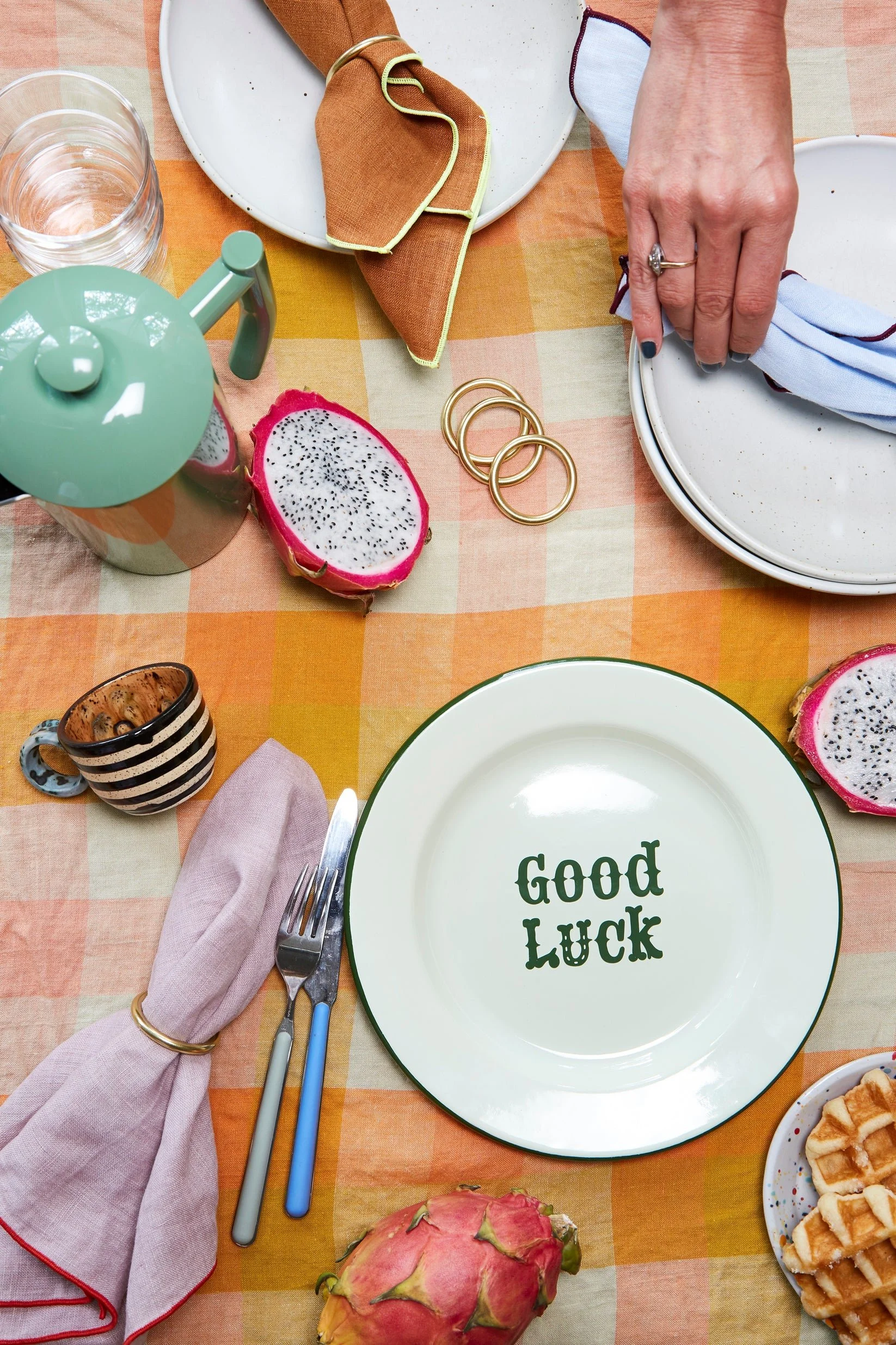 Table setting with a white plate with green rim that has the words "Good Luck" written on it, a fork and two blue-handled knives, a folded pink napkin, a watermelon, a dragon fruit, a teapot, a glass of water, a partially visible plate with waffles, a cups, and decorative gold rings on a colorful checkered tablecloth.