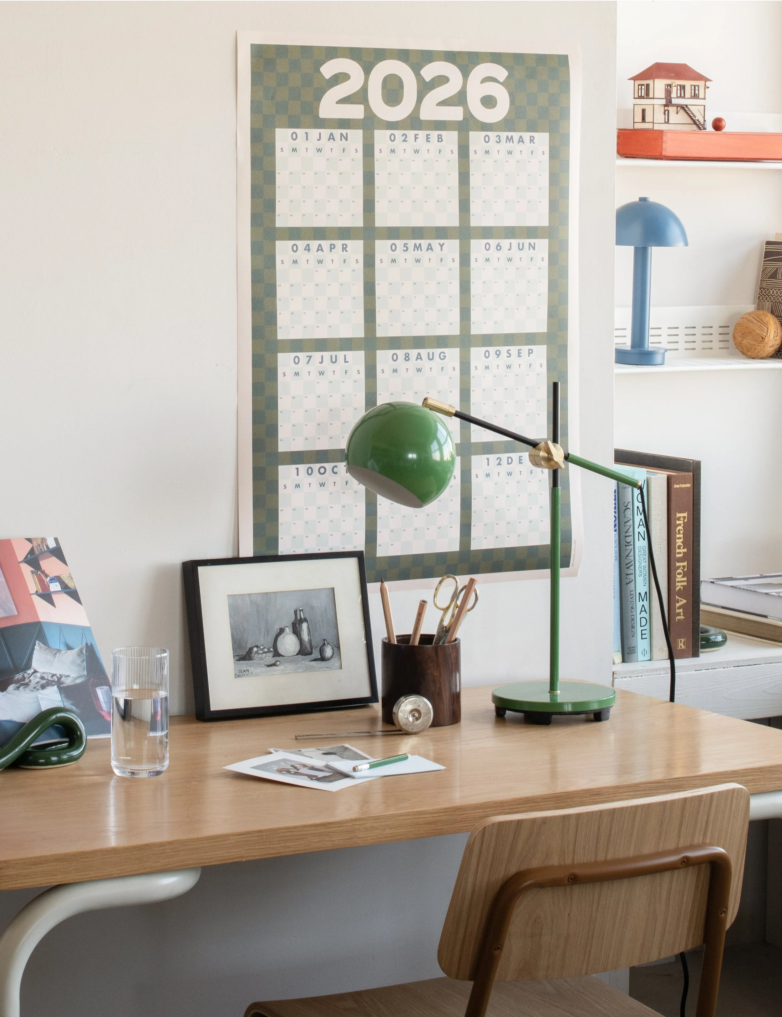 A home office workspace featuring a wooden desk, a green desk lamp, a glass of water, a black picture frame with a grayscale photo, a pencil holder with pencils, and a calendar on the wall showing months from January to December 2026. In the background, a blue lamp, books, and a small model house on a shelf are visible.