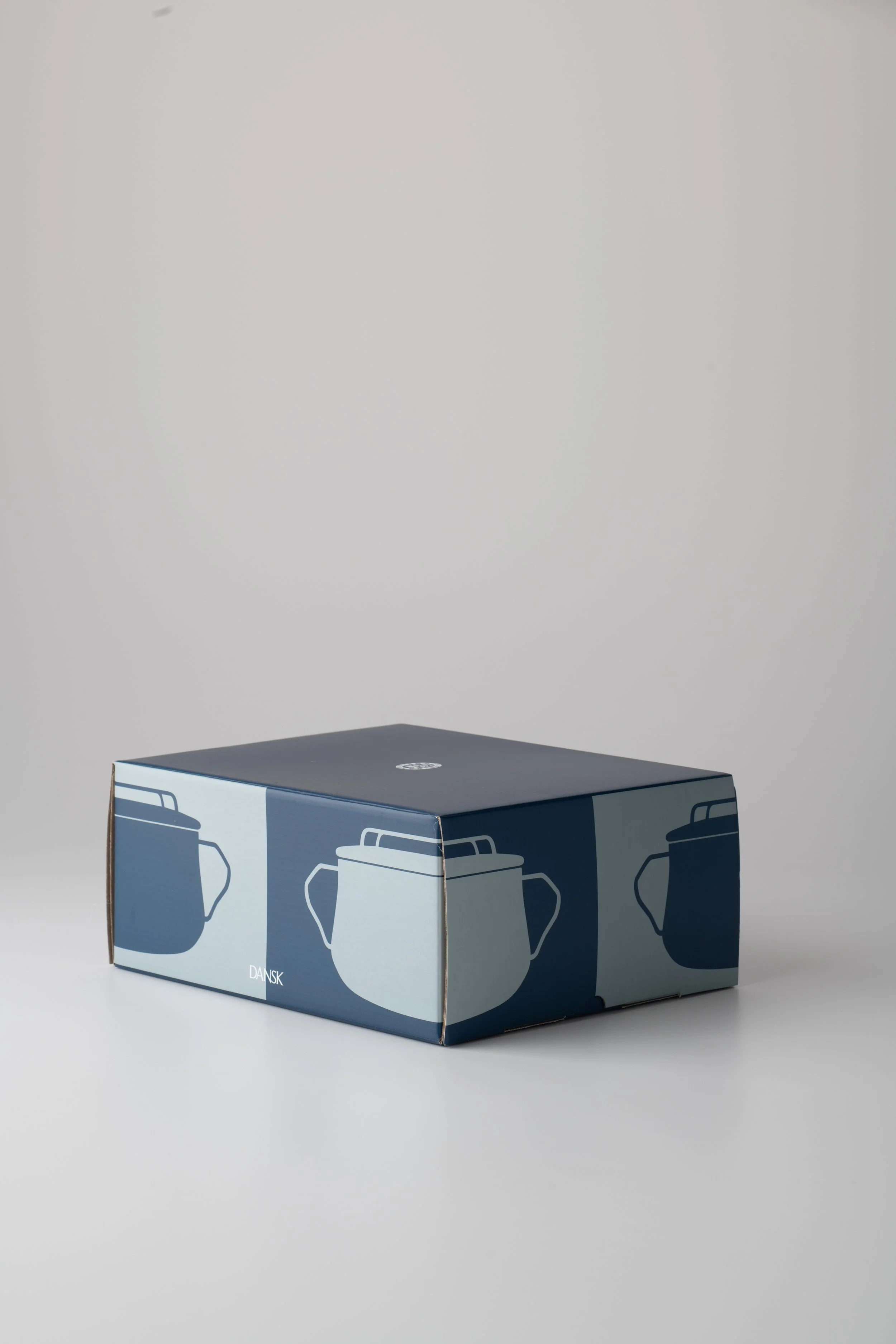 Blue cardboard box with repeating pattern of white and dark blue coffee mugs, placed on a white surface against a gray background. The word 'DANSK' is visible on the front of the box.