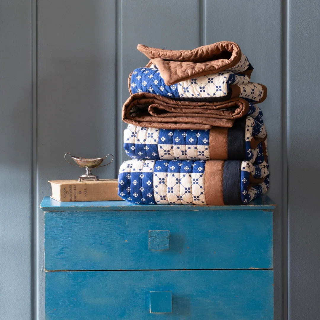 Stacked vintage quilts on a blue wooden dresser, with a small open book and a silver dish beside it, against a gray wall.