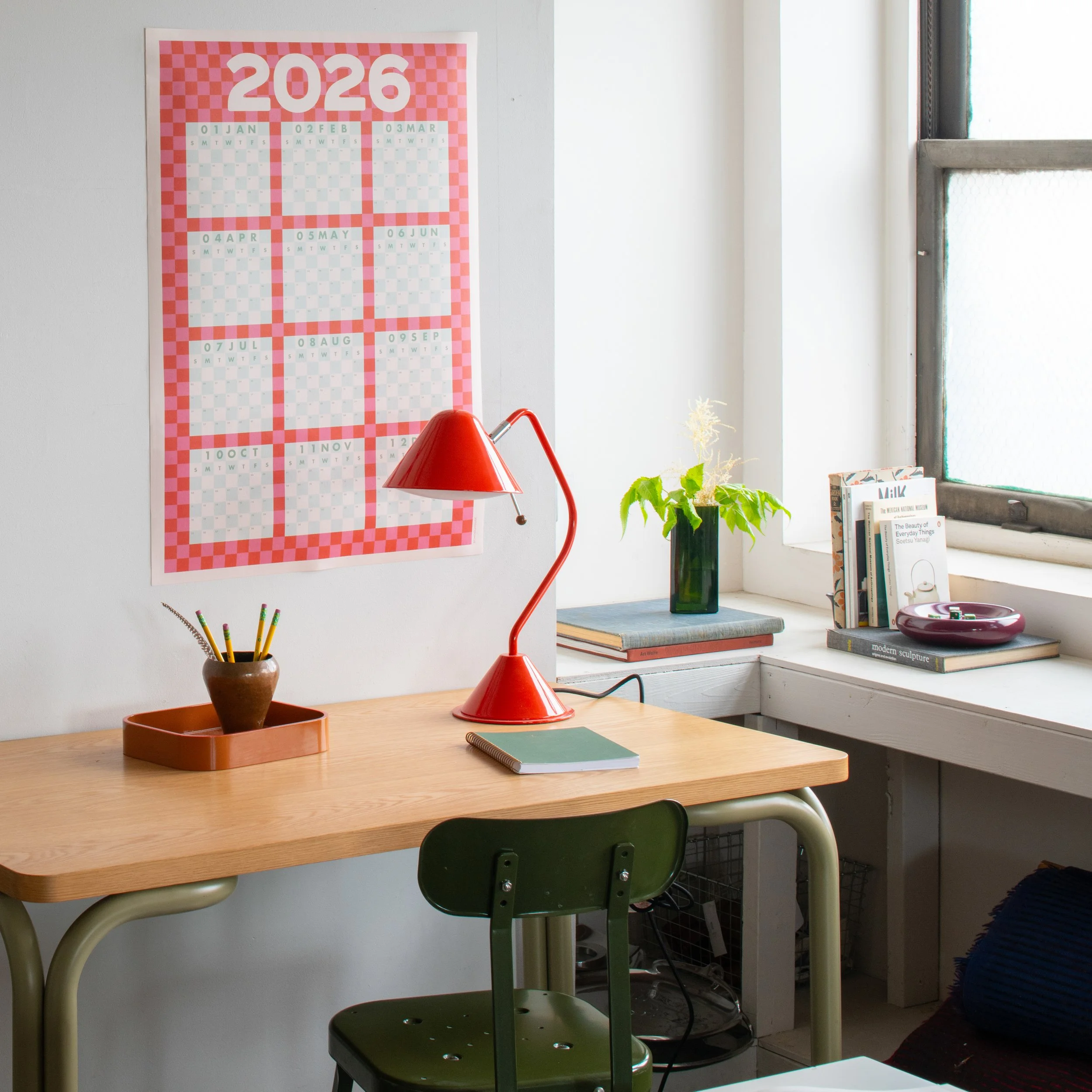 A desk with a brown pot holding colorful pencils, a red desk lamp, and several stacked books. Behind the desk is a window with a wide windowsill and a white wall with a pink and white calendar on it.