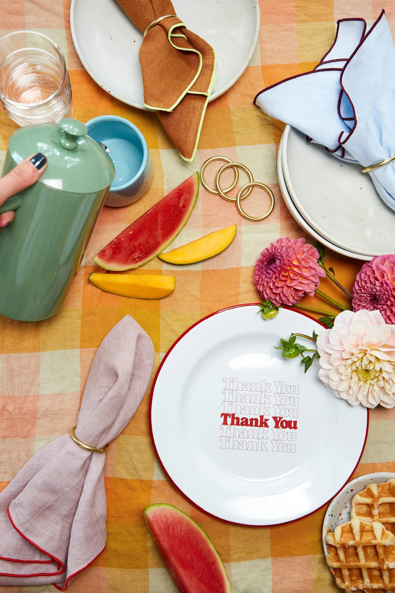 Table setting with watermelon slices, a plate with a "Thank You" message surrounded by flowers, a napkin, and glassware on a checkered tablecloth.