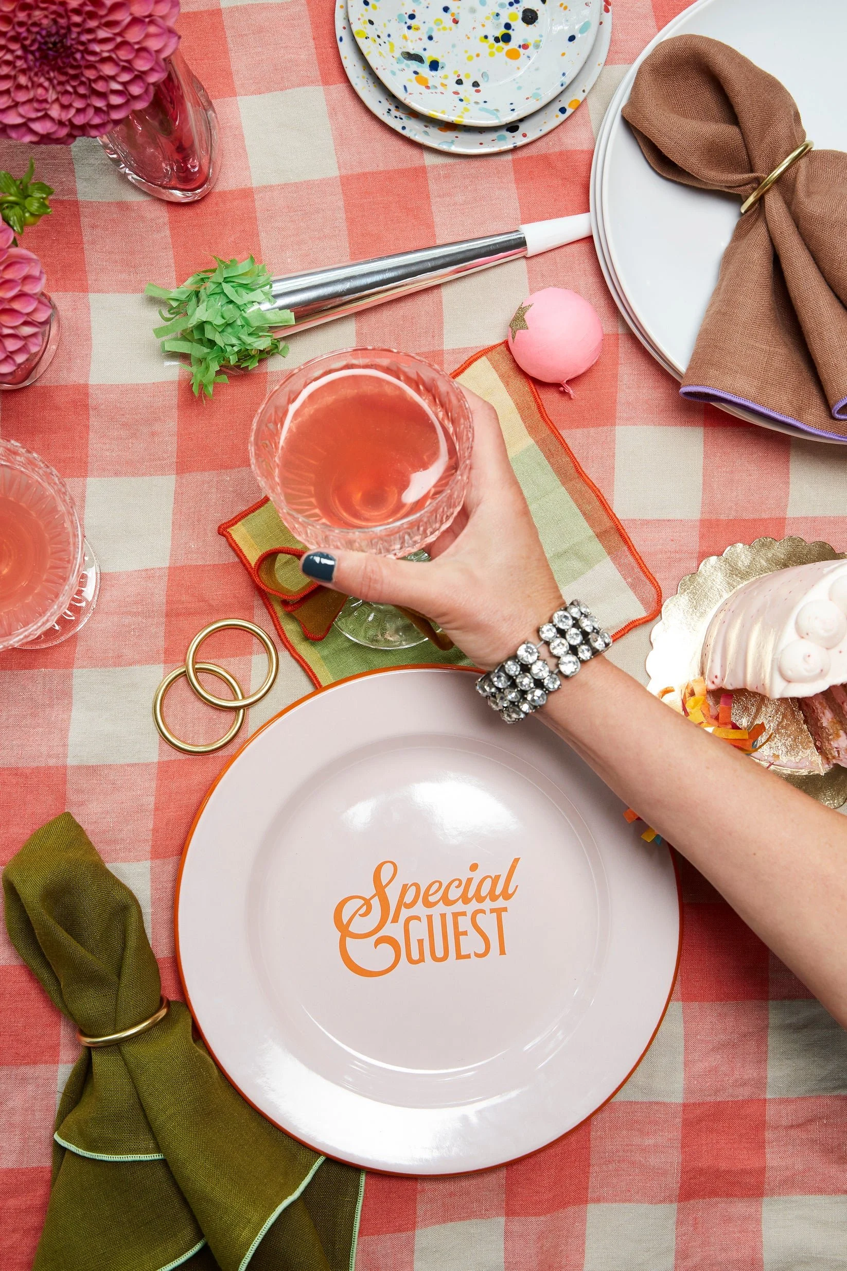Table setting with a pink and beige checkered tablecloth, decorated with pink flowers, a green fabric napkin, gold rings, utensils, plates with colorful splatters, a glass of pink drink, a plate with the words "Special Guest" in orange, and various party decorations.