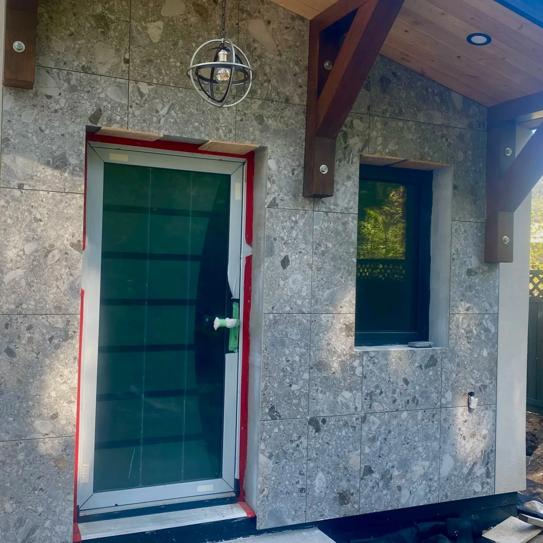 A glass door installation is being done on a stone wall with visible tools and supplies. The door frame is surrounded by masking tape, and cleaning supplies are placed on the bottom of the door. The scene appears to be in a construction or renovation phase.