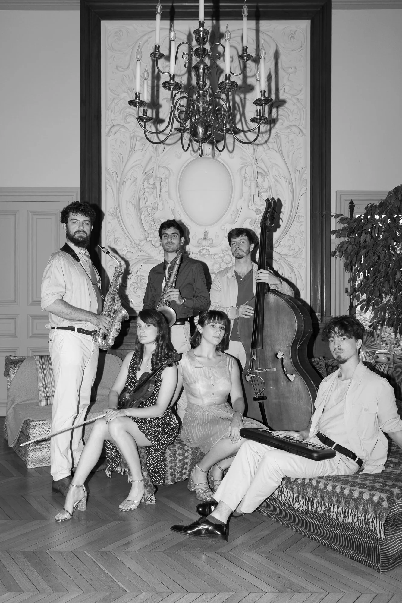Un groupe de musiciens posant dans une salle élégante avec un chandelier au plafond. Il y a trois hommes et trois femmes, certains avec des instruments tels qu'un violon, un contrebasse, un clavier et des saxophones.