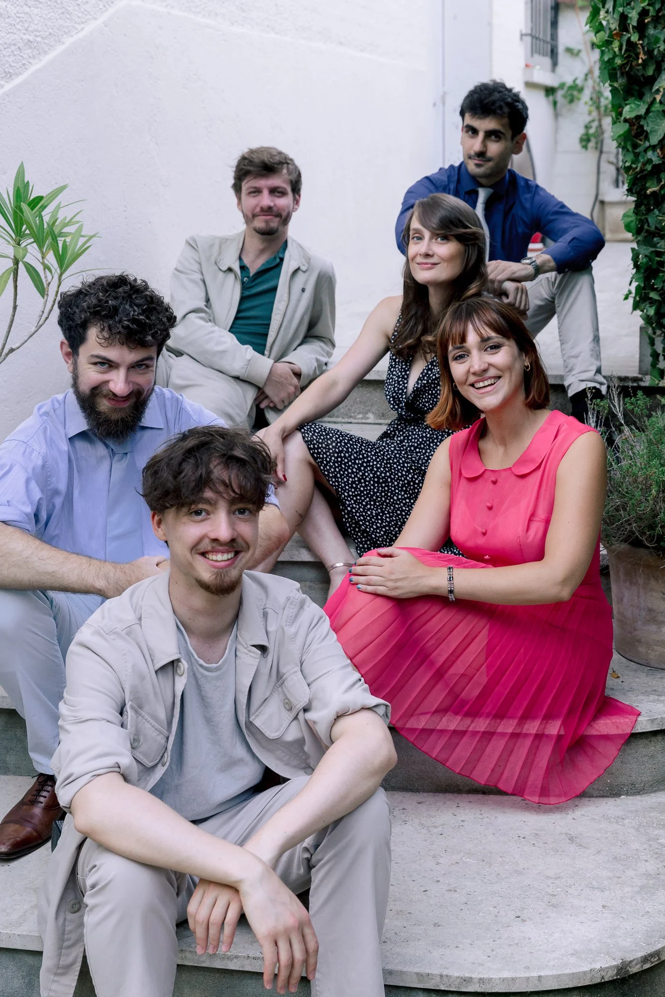 Un groupe de six jeunes adultes posant pour une photo sur un escalier extérieur avec plantes et mur blanc en arrière-plan.