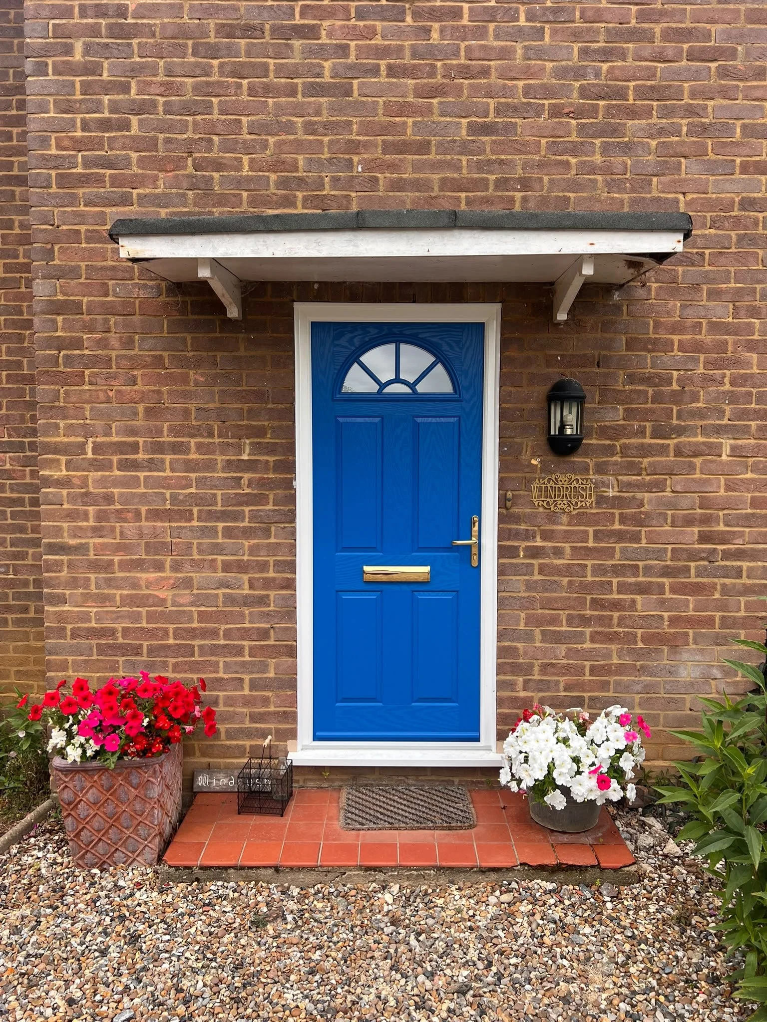 Front door of a house with a bright blue door, brick wall, a small porch with a black outdoor light, and colorful flower pots with red, white, and pink flowers on either side of the door.