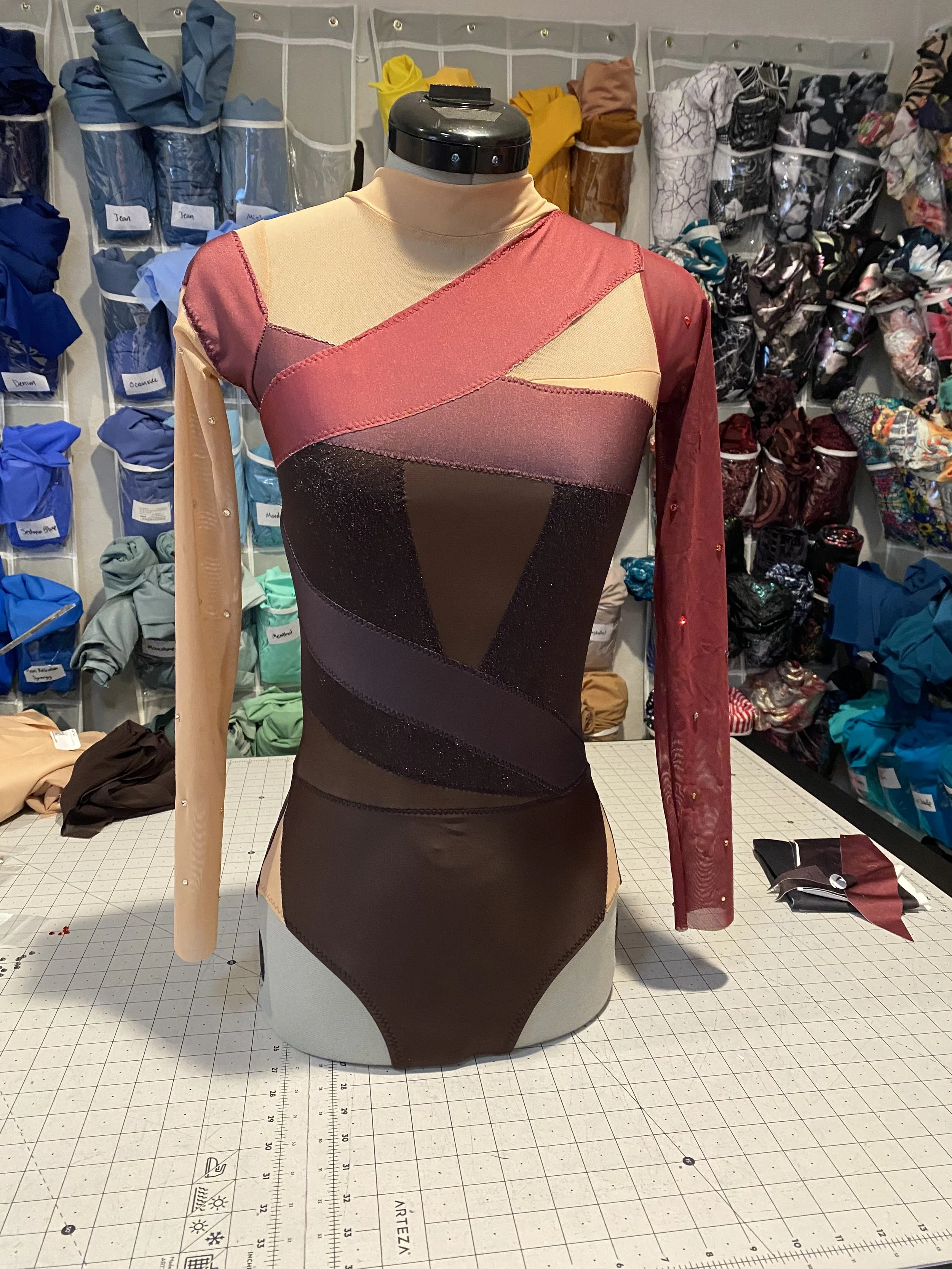 Close-up of a dress form wearing a multicolored, geometric-patterned dress with diagonal paneling in shades of pink, red, black, and beige, on a cutting table in a fabric store or workshop. In the background are fabric swatches and shelves filled with various folded and packaged fabrics.