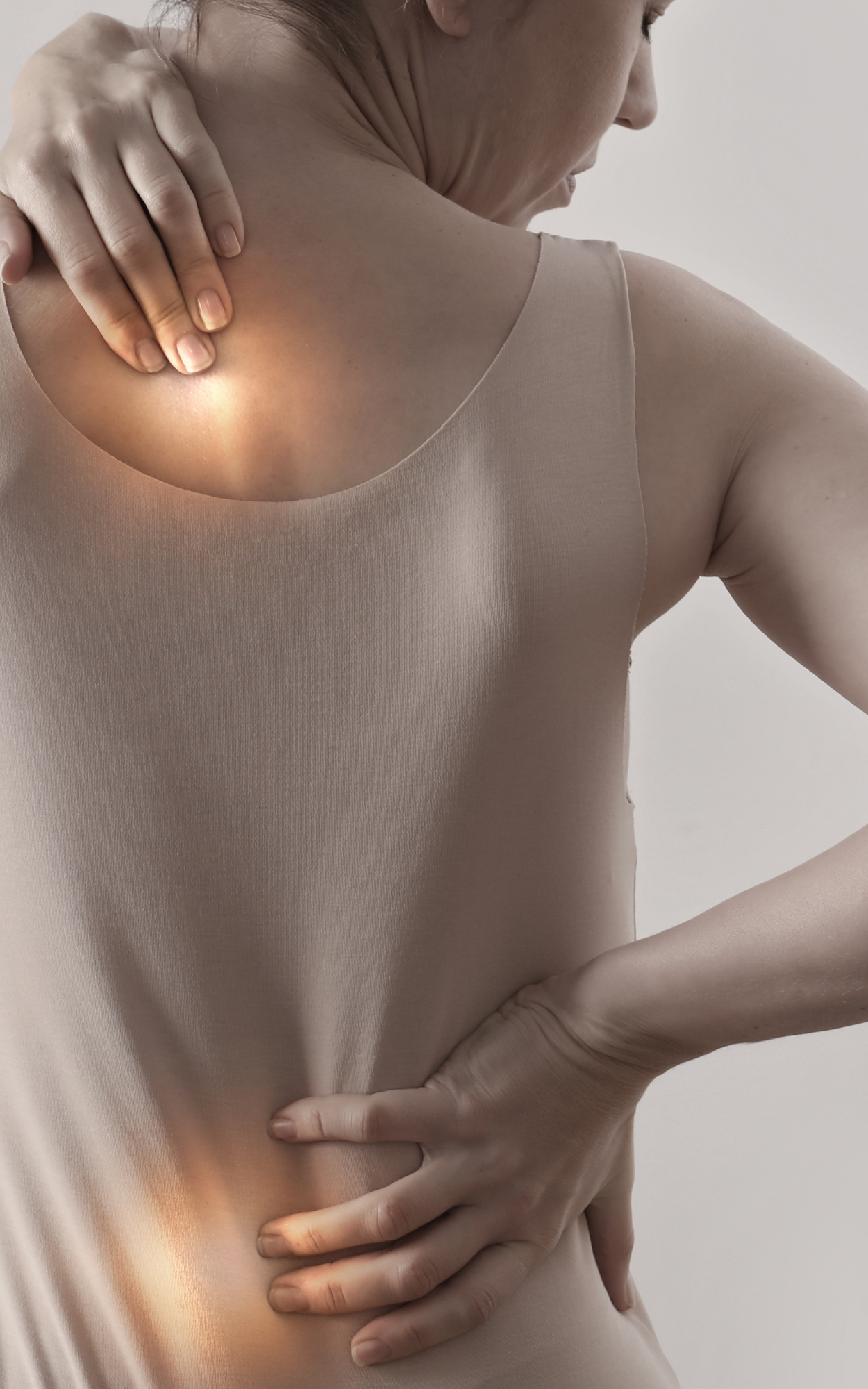 A person with fair skin and brown hair, wearing a beige sleeveless top, is holding their upper back and lower back with both hands, pressing against soreness or pain.