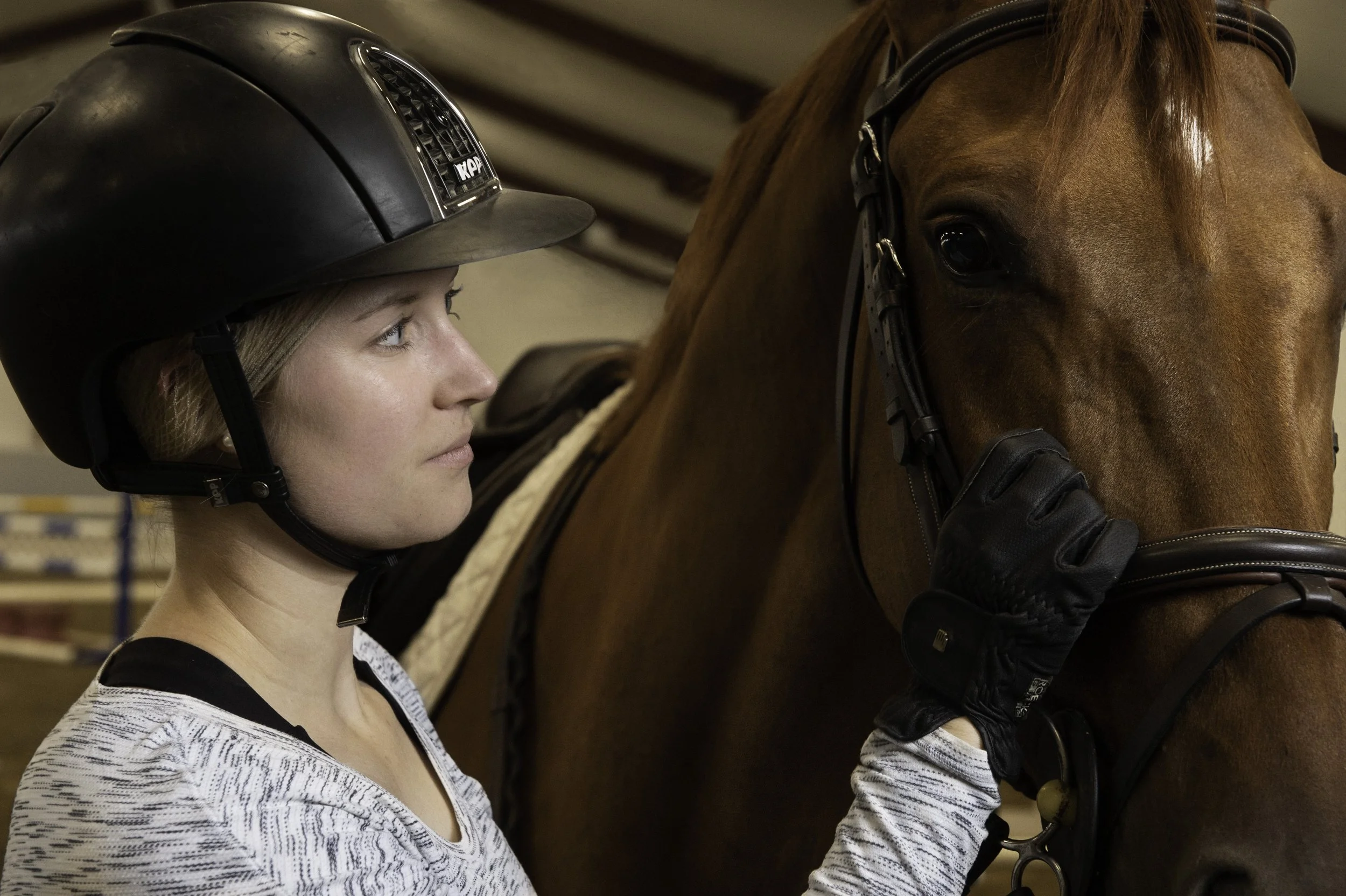 A woman wearing a black riding helmet gently touching the face of a brown horse in an indoor stable.