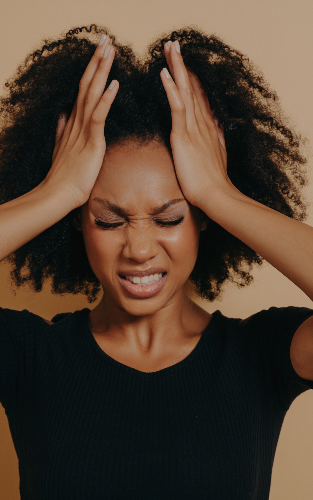 A woman with curly hair holds her head with both hands, her eyes are closed, and she appears to be experiencing discomfort or stress.