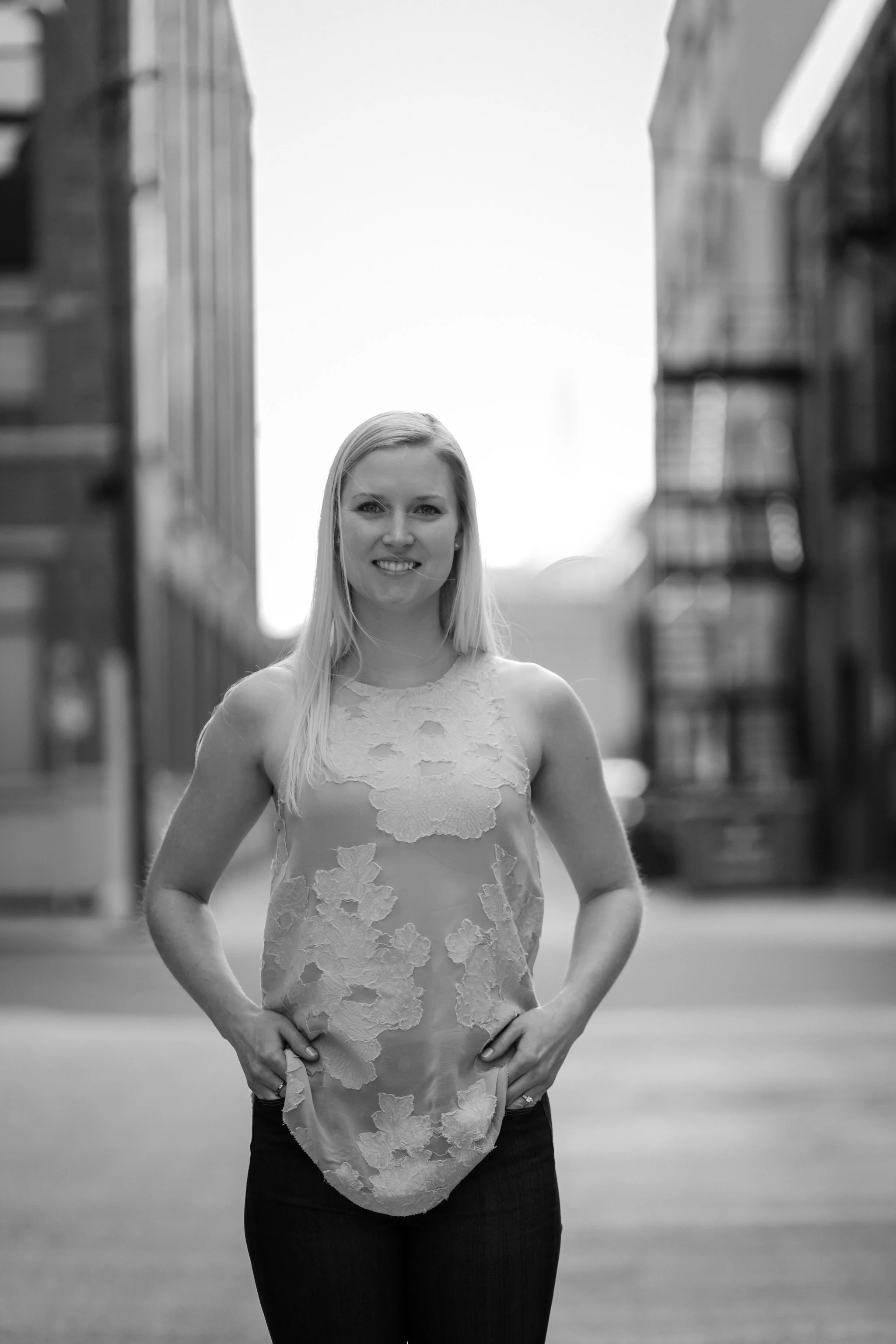 Black and white photo of a woman smiling, standing in front of a brick building, with her hands on her hips, wearing a lace dress and dark jeans.