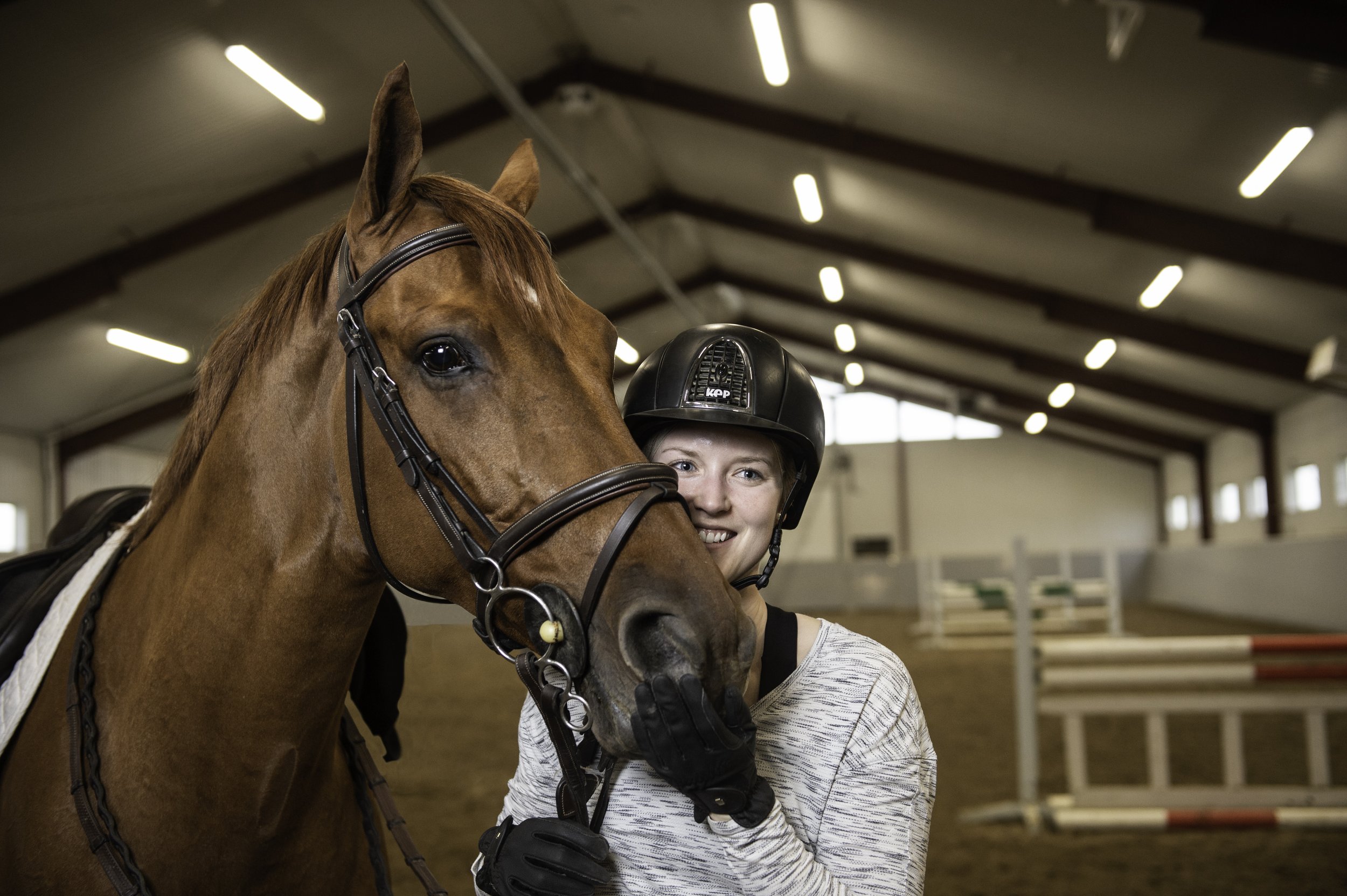 A young woman smiling and holding a brown horse in an indoor riding arena.