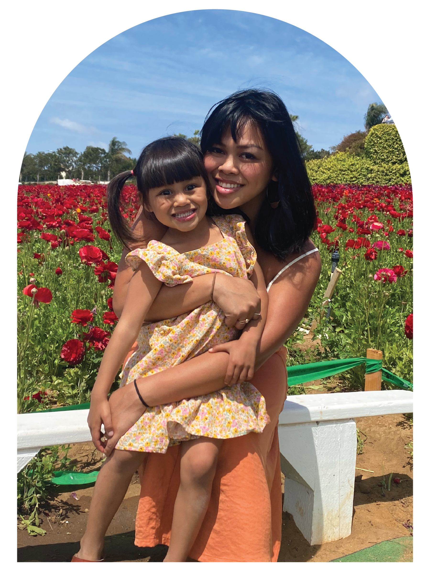 A woman with black hair holding a young girl with dark hair styled in a ponytail, standing in a field of red flowers with trees and blue sky in the background.