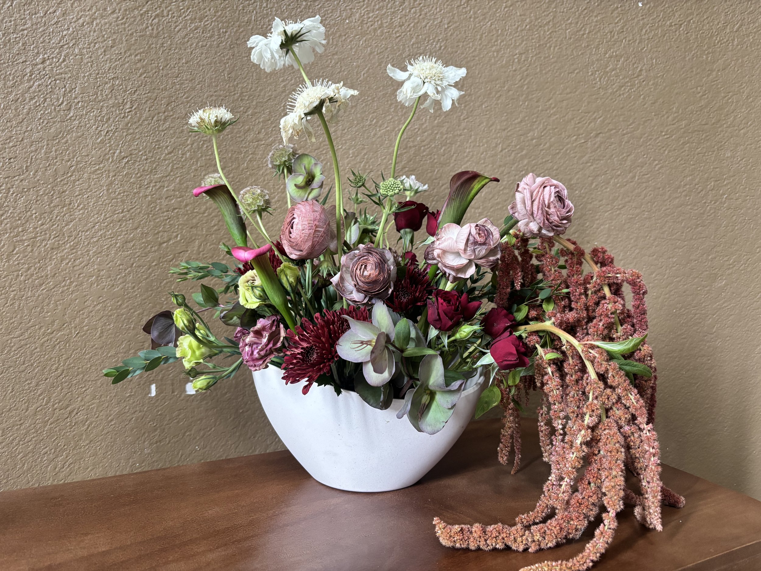 A floral arrangement in a white vase, featuring white, pink, and burgundy flowers with greenery on a wooden surface against a beige wall.