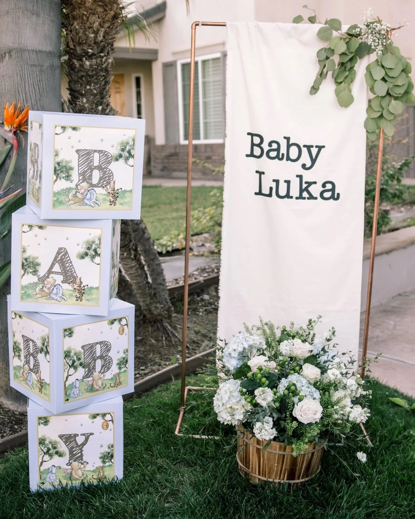Decorative outdoor setup for a baby named Luka, featuring a white banner with black text, a floral arrangement with white flowers in a wooden basket, and illuminated boxes with illustrations of baby animals and a large letter 'B'.