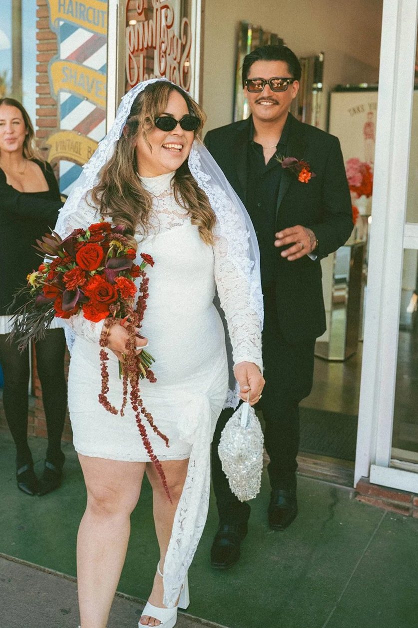 A woman in a white lace dress and veil holding a bouquet of red and dark purple flowers, smiling and wearing sunglasses. A man in a black suit with sunglasses and a boutonniere stands behind her. They are outside a shop with signage for haircuts and shaves.