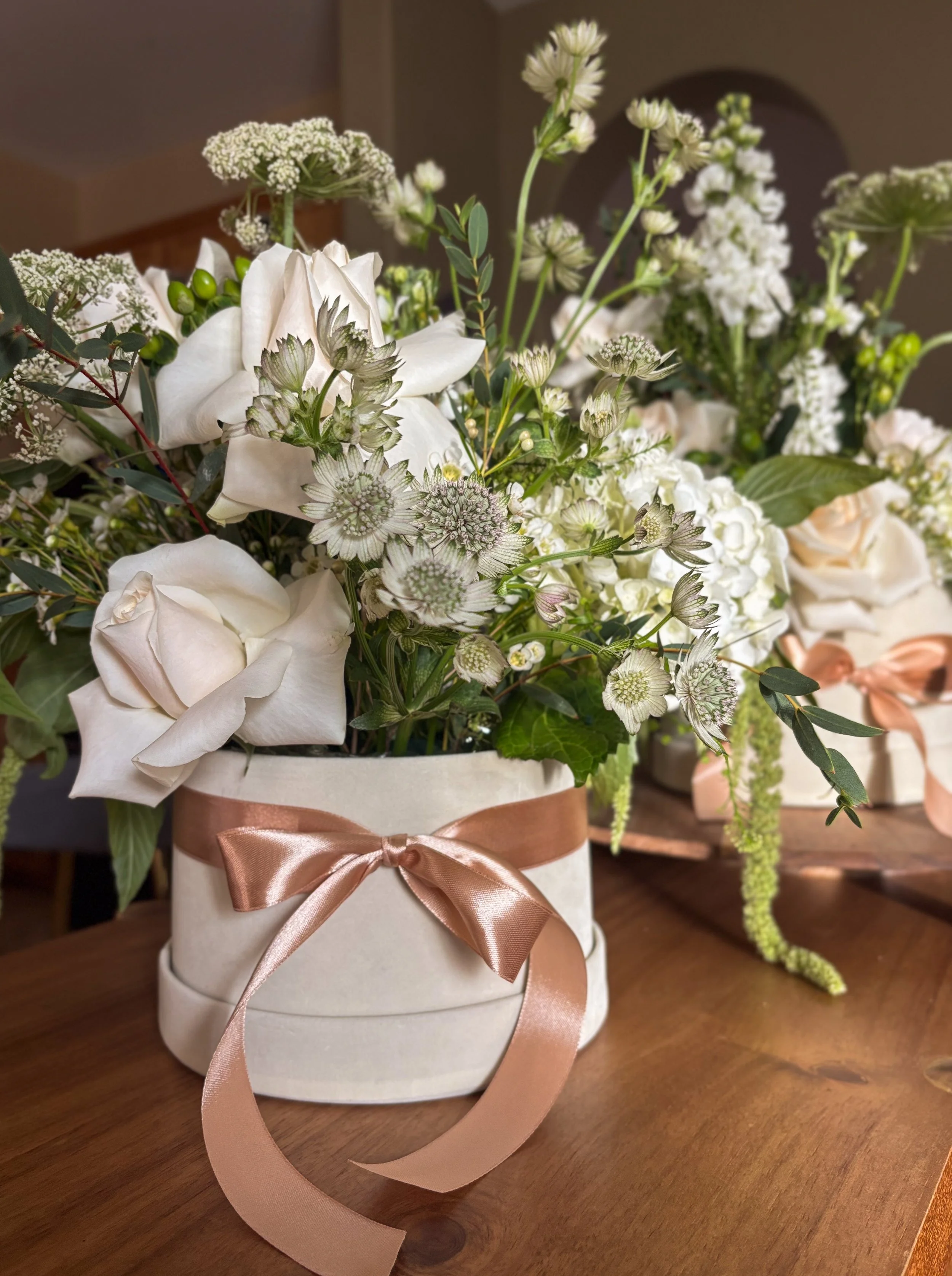 A bouquet of white flowers with green leaves in a white ceramic vase decorated with a pink satin ribbon tied in a bow, placed on a wooden surface.