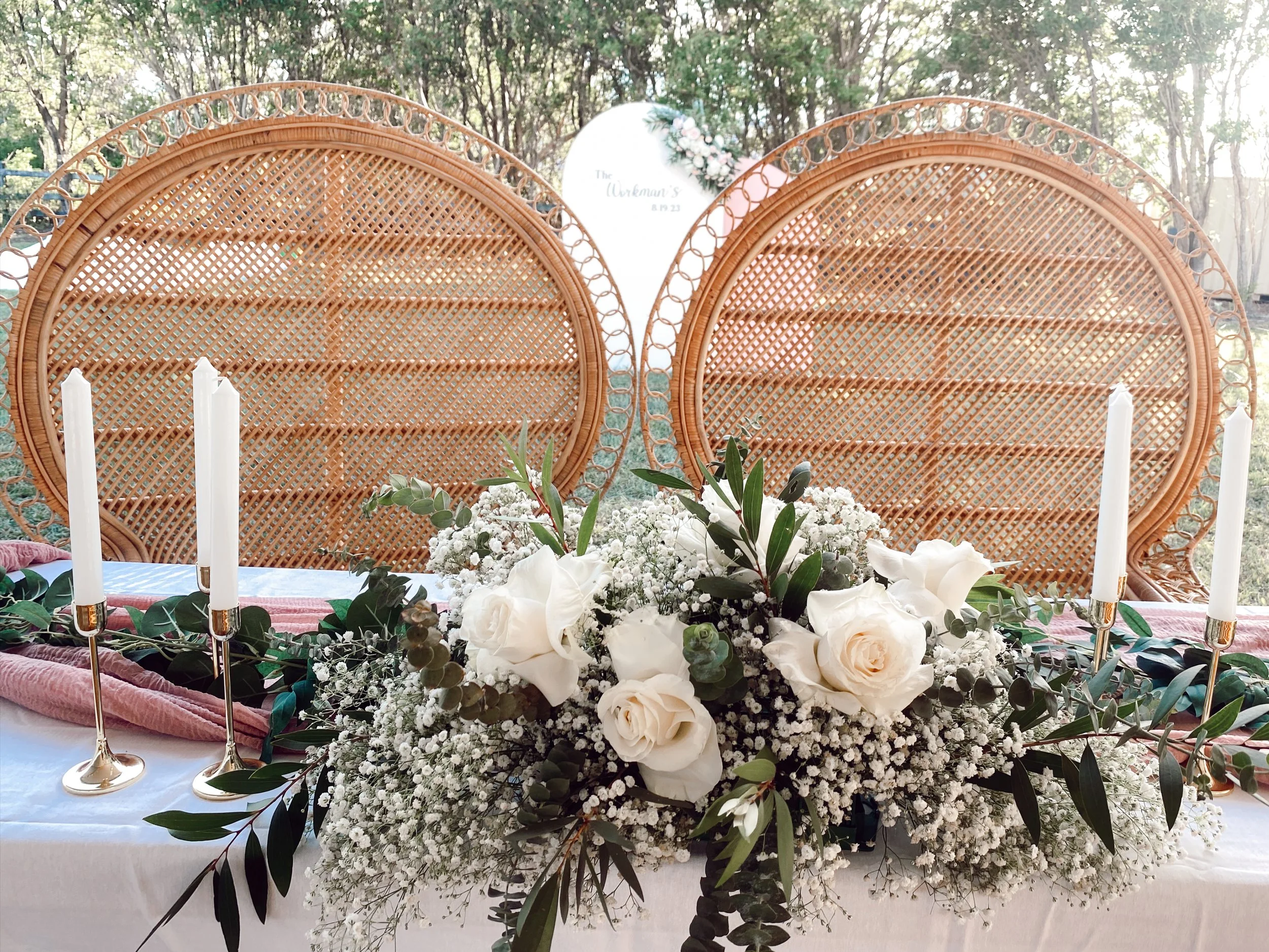 Wedding ceremony decor with a centerpiece of white roses, baby's breath, and greenery, surrounded by four tall white candles on a table, and rattan chairs and trees in the background.