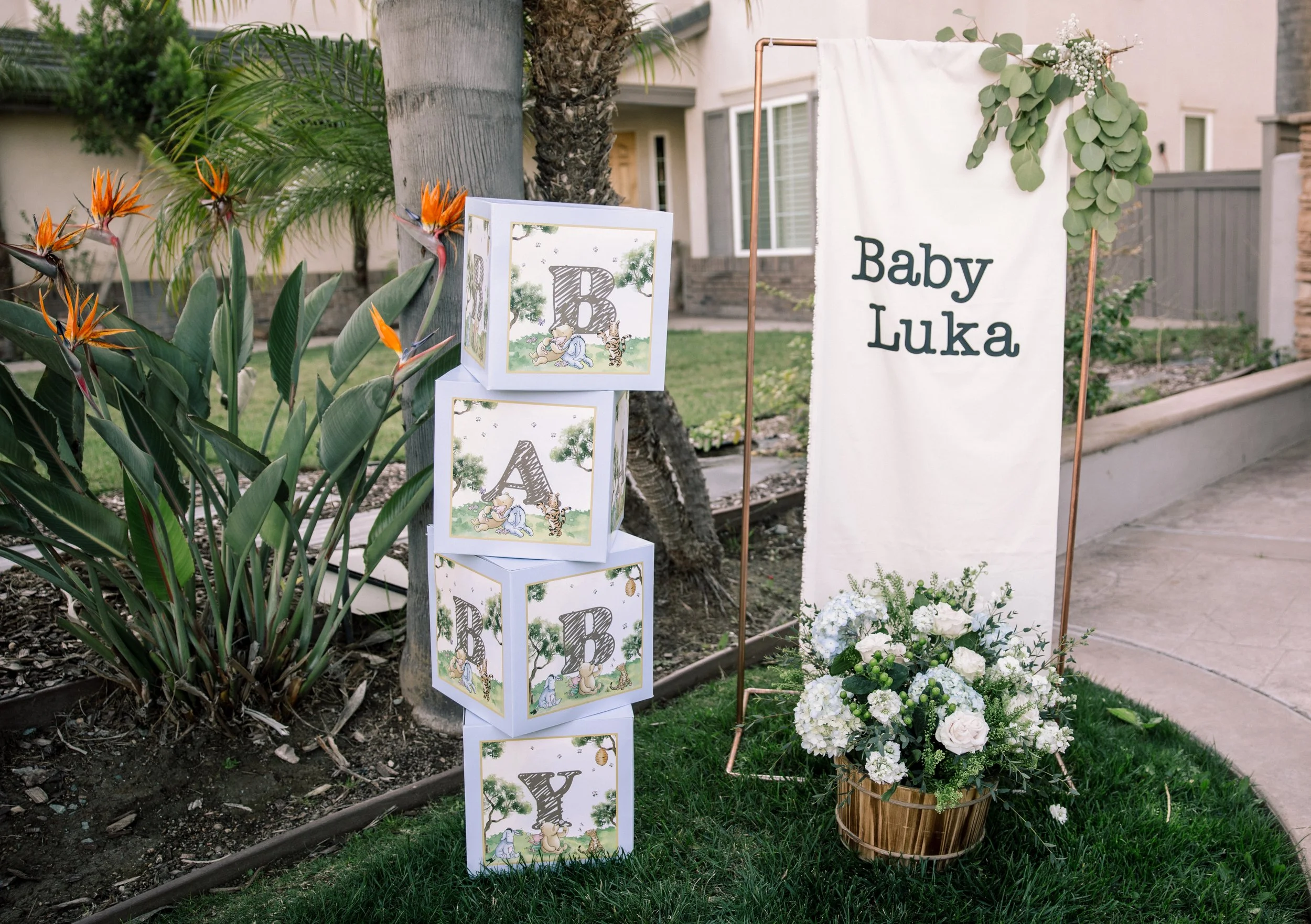 Outdoor nursery or baby shower decor with a white sign that says 'Baby Luka', decorative boxes with woodland animal and alphabet themed designs, and a flower arrangement in a basket, surrounded by greenery and plants.