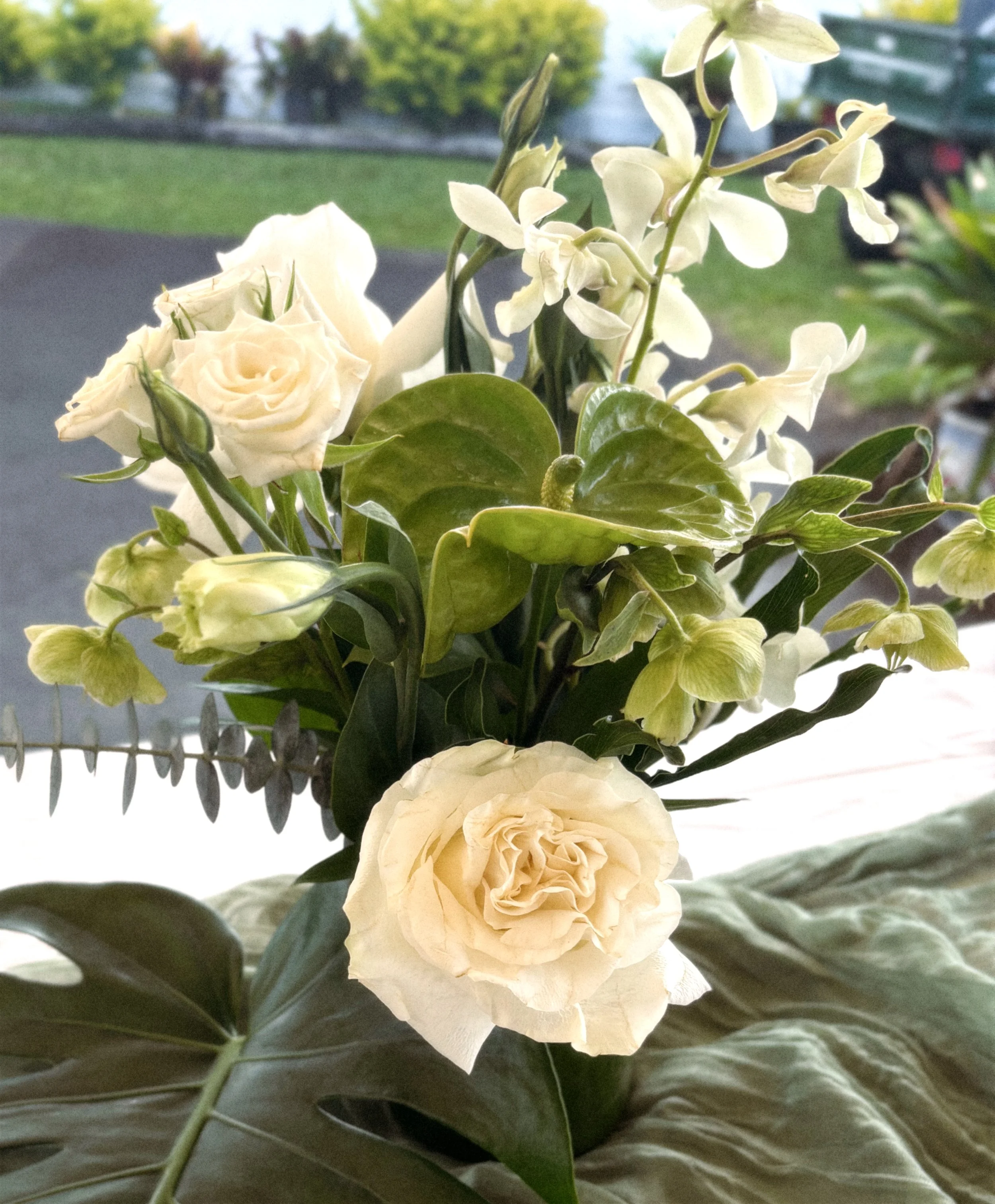 A bouquet of white roses and white orchids with green leaves, positioned against a window with a blurred outdoor background of trees and grass.
