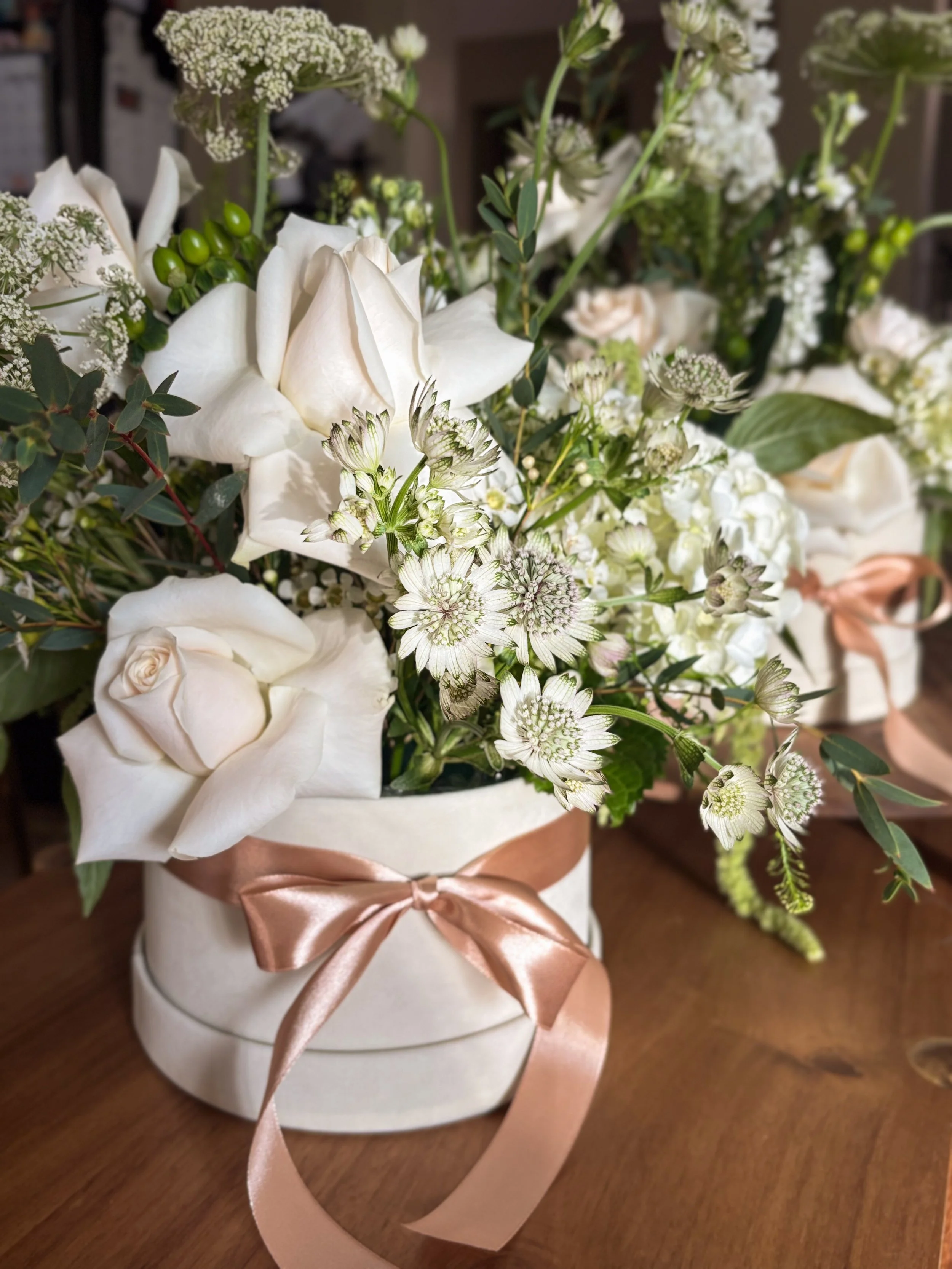 White flower bouquet with roses and greenery in a white vase adorned with a pink satin ribbon on a wooden surface.