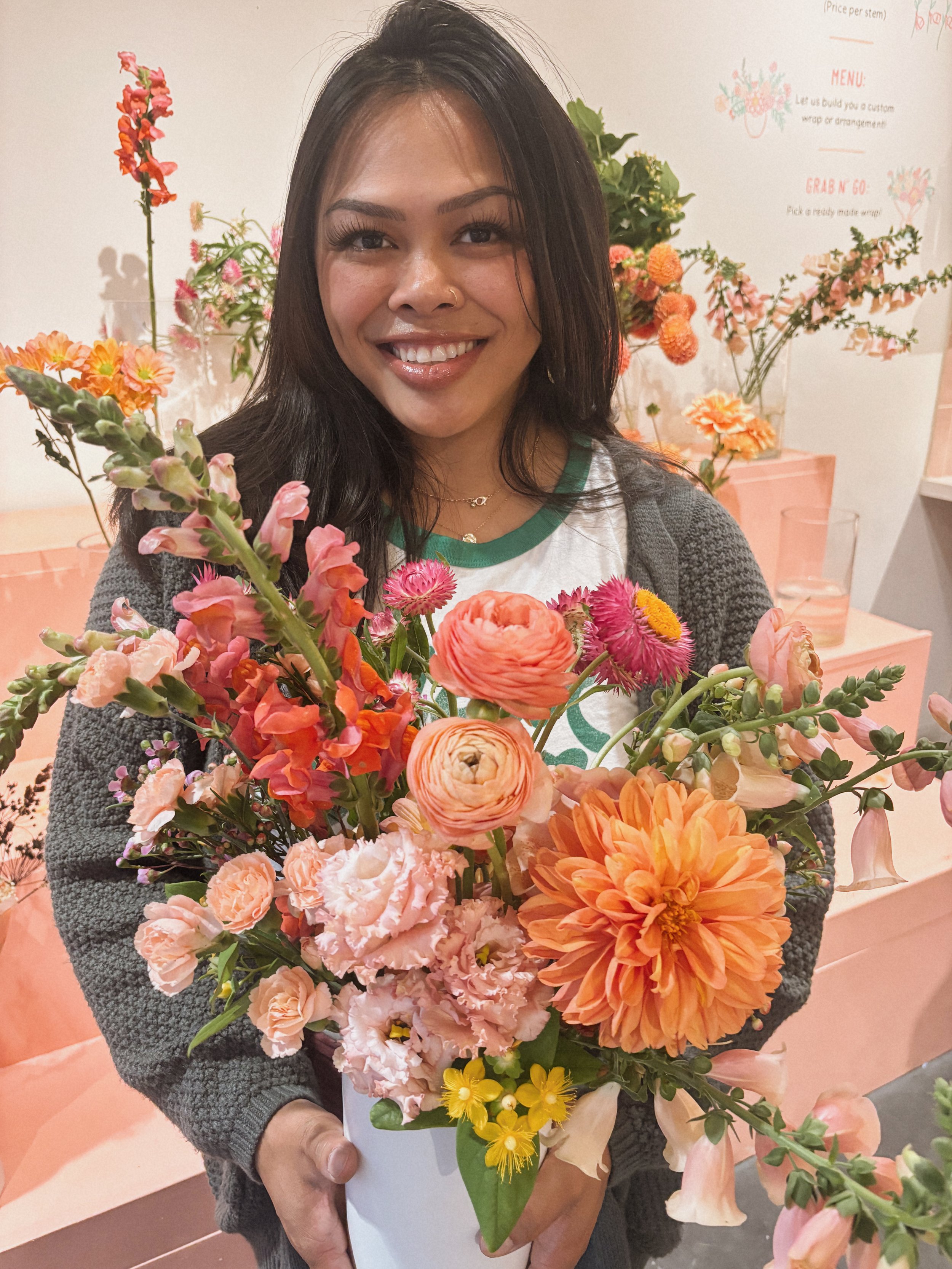 A woman smiling and holding a bouquet of multicolored flowers, standing in front of a white wall with floral decorations.