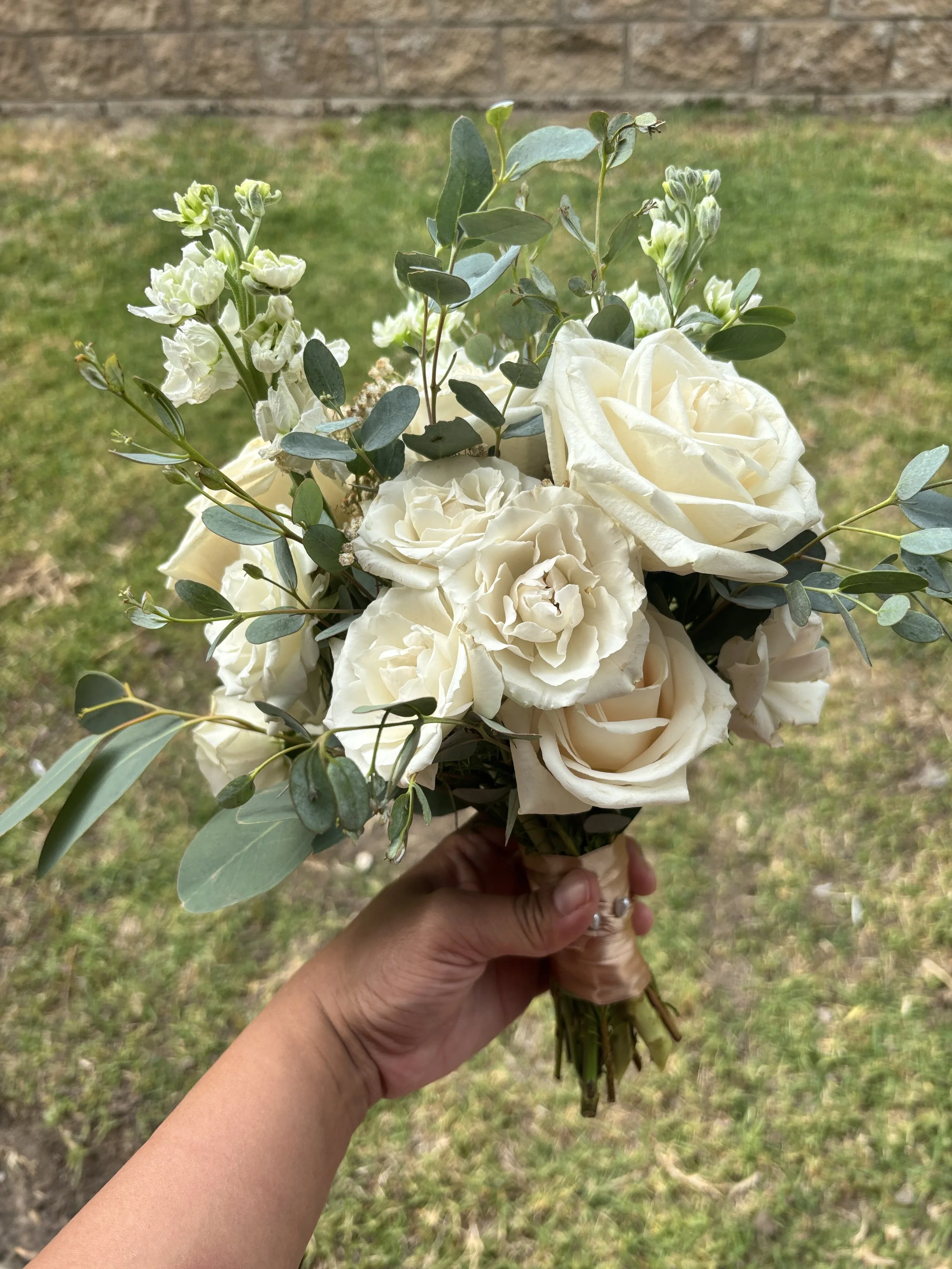 A person holding a bouquet of white roses and green eucalyptus leaves outdoors, with a grass and brick wall background.
