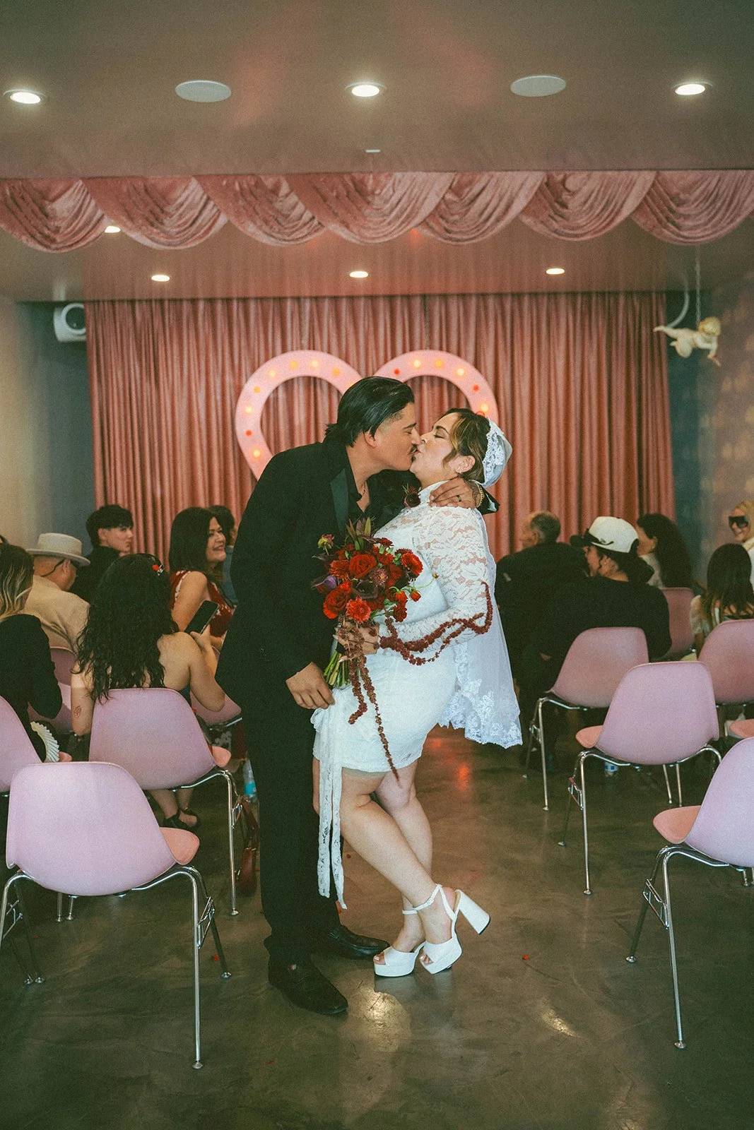 A couple kisses at a wedding ceremony. The woman wears a white lace dress and high heels, holding a bouquet of red and purple flowers. The man is in a black suit. Guests sit on pink chairs in the background, with pink curtains and a heart-shaped neon