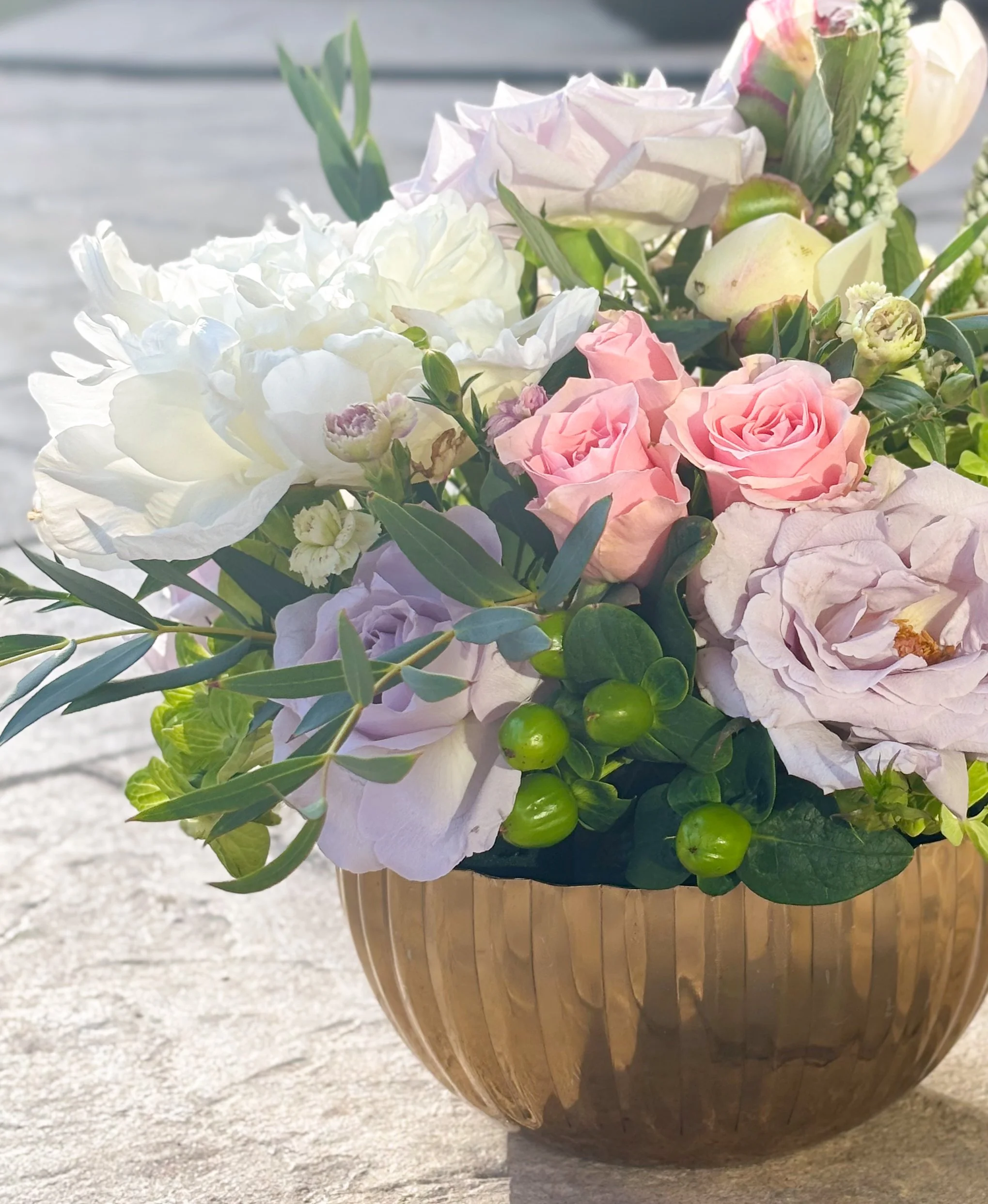 A wooden vase filled with white, pink, and cream-colored roses, green berries, and various green leafy foliage, placed on a concrete surface.