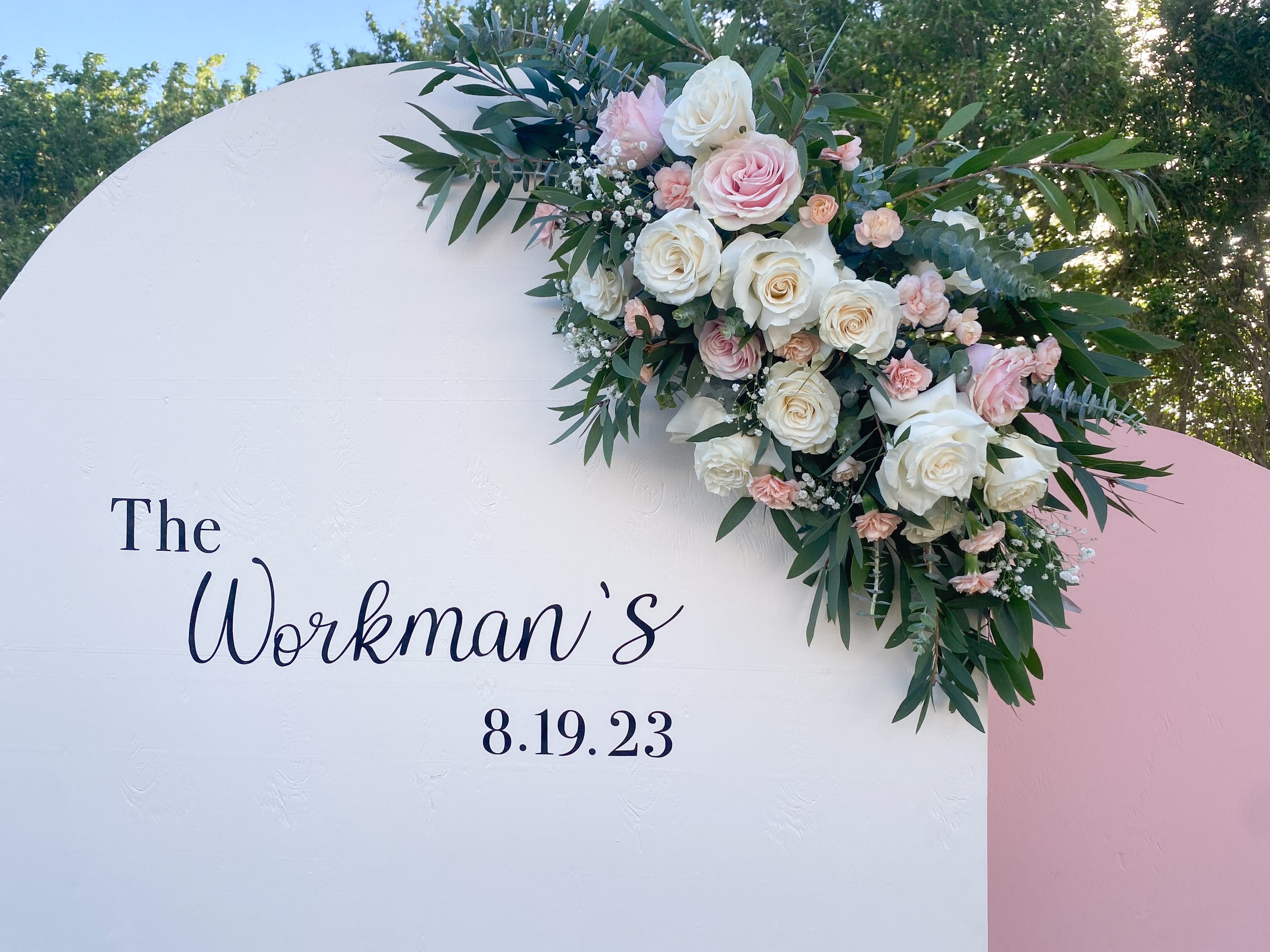 A floral wedding arrangement with white and pink roses, baby's breath, and green leaves on a white sign at an outdoor ceremony.